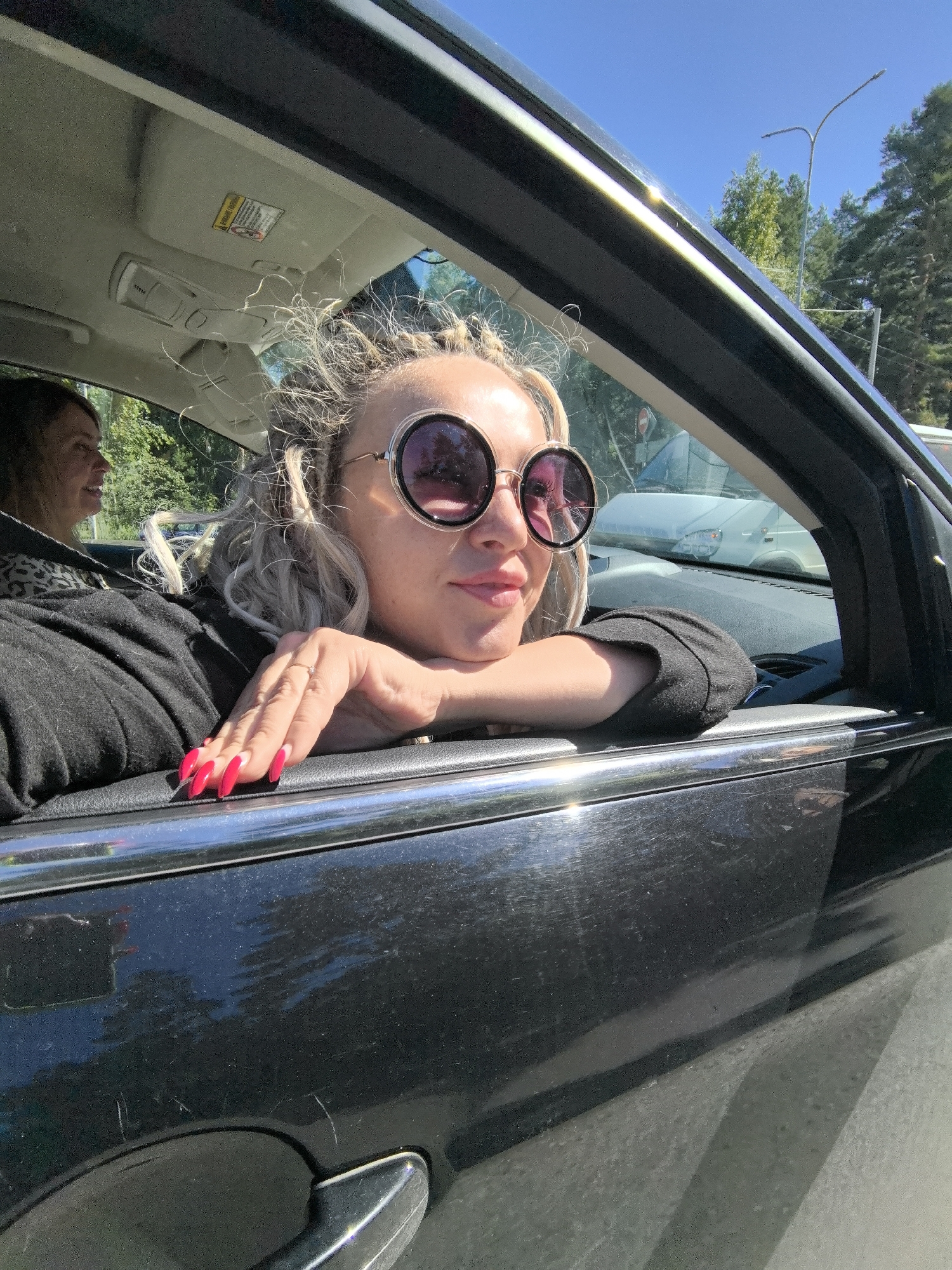 A woman is leaning out of a car window with her arm resting on the car door. She is wearing large, round sunglasses and her hair is blonde and styled in braids. Her fingernails are painted bright pink. A second woman is visible in the background, sitting in the passenger seat. The car appears to be black. The background shows trees and a street light, suggesting an outdoor, possibly urban setting. The image is taken from the perspective of someone outside the car, looking in. The location is specified as Chaikovsky, Russia.