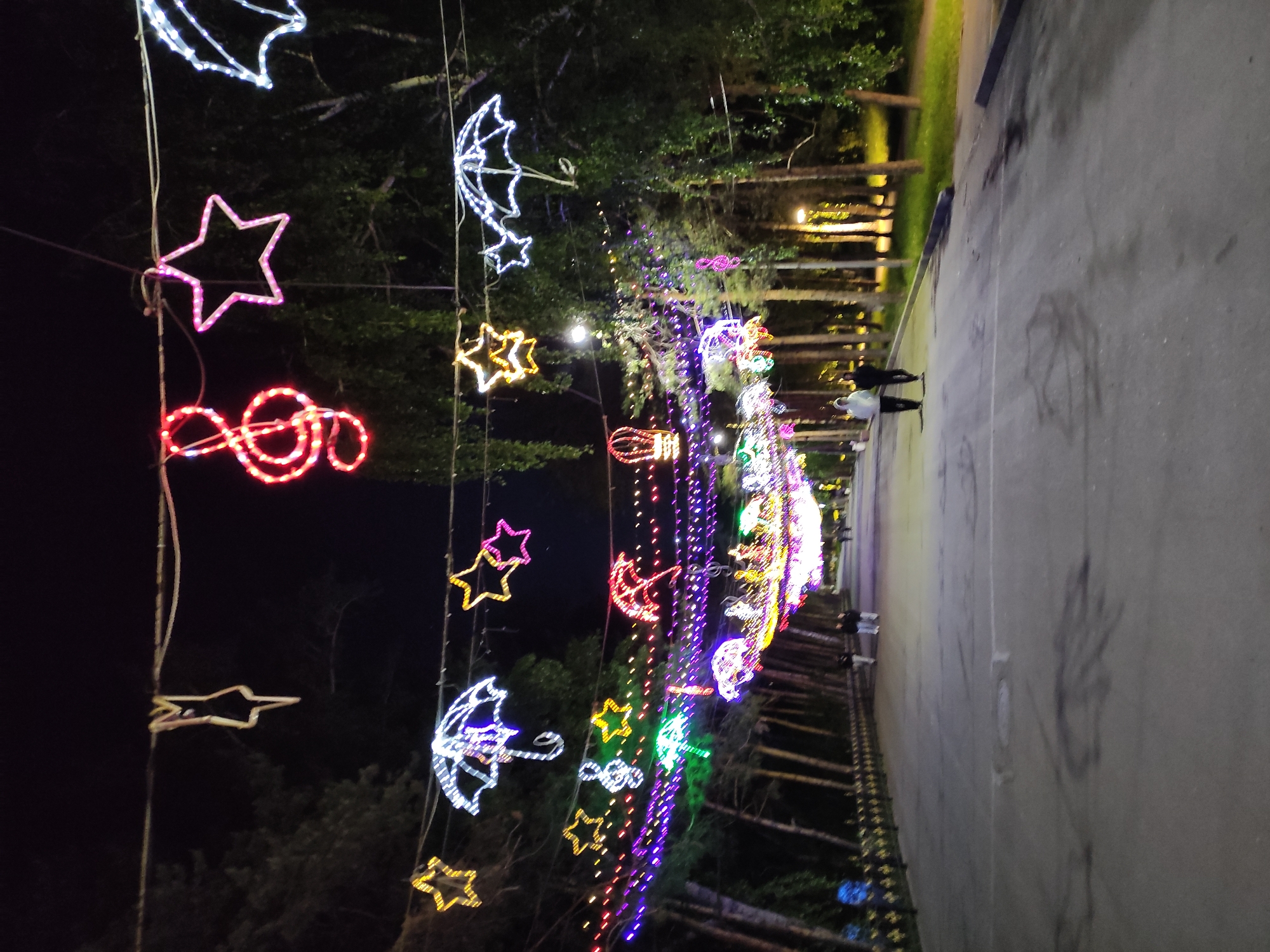 The image shows a nighttime view of a pedestrian walkway decorated with lighted figures of stars, umbrellas, and musical notes. These figures are strung along overhead wires. On the right side of the image is a paved walkway, and beyond the lights are trees and possibly parkland. Two people are walking in the distance along the walkway. The location is Pavlodar, Kazakhstan.