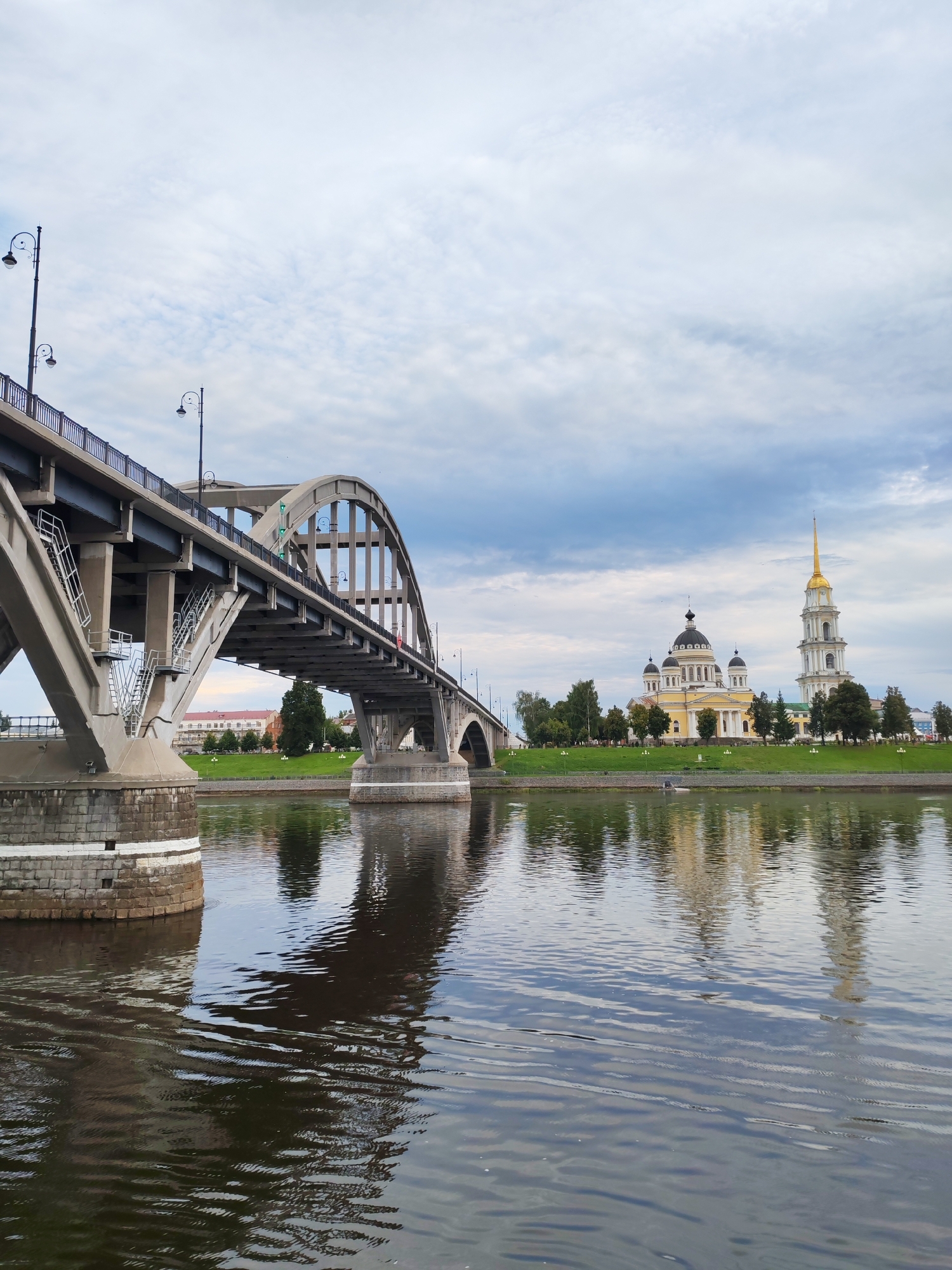 The image captures a scenic view of a bridge spanning a body of water in Bol'shie Mkhi, Russia. The bridge, constructed of concrete and steel, features a prominent arch design and is supported by massive stone piers. Several streetlights are visible along the bridge's railing. On the opposite bank of the river, a distinct yellow cathedral with multiple domes and a tall, golden spire dominates the skyline. The cathedral is surrounded by lush green grass, trees, and other buildings. The water in the foreground reflects the bridge, the cathedral, and the cloudy sky, creating a slightly distorted but captivating mirrored image. The overall atmosphere is calm and serene, suggesting a peaceful day in this Russian city.