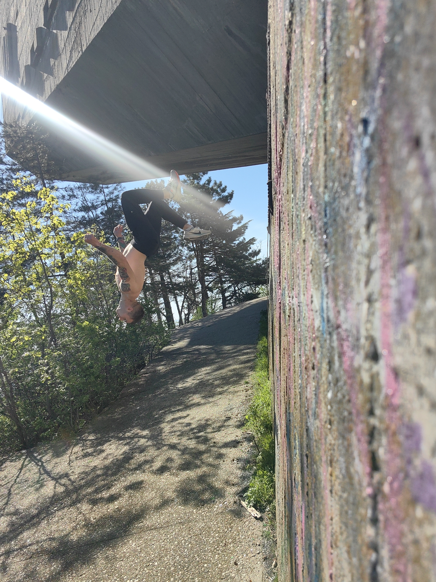 A man is performing a backflip near the Asparuhov Bridge in Varna, Bulgaria. He is wearing black pants and white sneakers. He is shirtless and has tattoos on his torso and arms. The background includes trees, a road, and the bridge structure. A concrete wall is in the foreground, adorned with various colors of graffiti. The lighting is bright, with the sun visible in the upper left corner. The overall scene captures an athletic activity taking place in an urban environment.