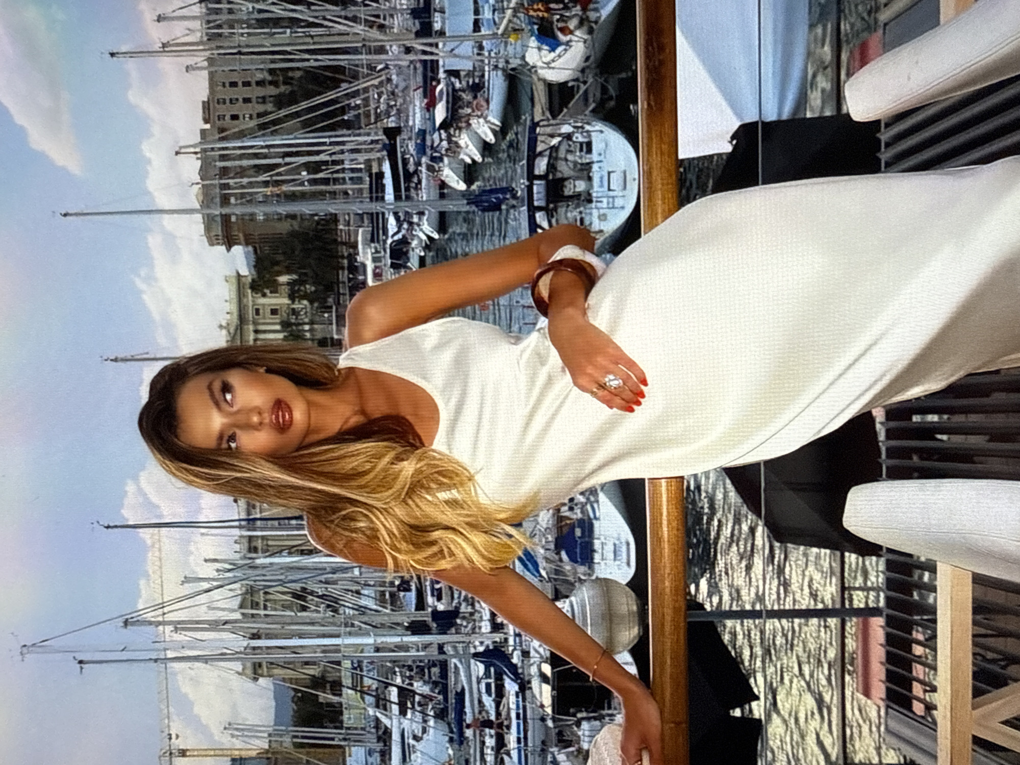 The image shows a woman in a white dress standing on a balcony overlooking a harbor filled with sailboats. She has long blonde hair and is wearing jewelry. The background features buildings and a construction crane. The water reflects the sky and boats.