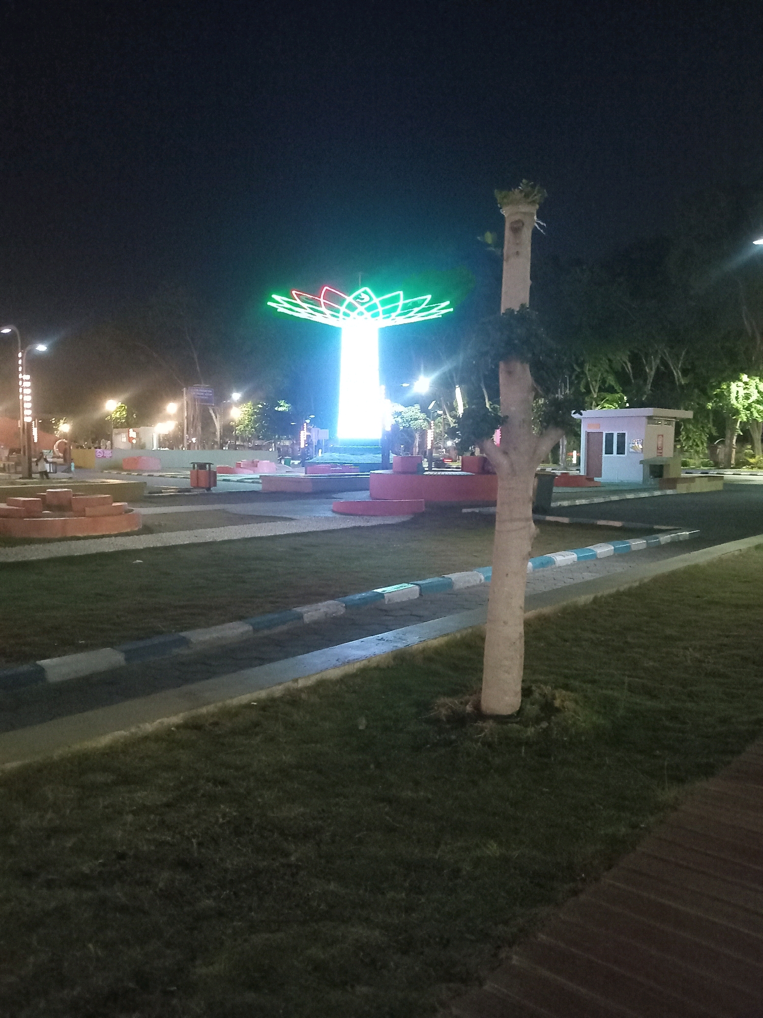 The image shows a night view of Taman Abhinaya in Sidoarjo, Indonesia. A prominent lotus-shaped light installation, glowing in green, pink, and white, stands in the center of the park. In the foreground, a narrow tree trunk with some foliage at its top stands beside a grassy area. The park features paved pathways with decorative borders of alternating white and blue blocks, as well as benches and some small structures. In the background, more trees and various lights from street lamps and buildings are visible. There are no people clearly visible in the image.