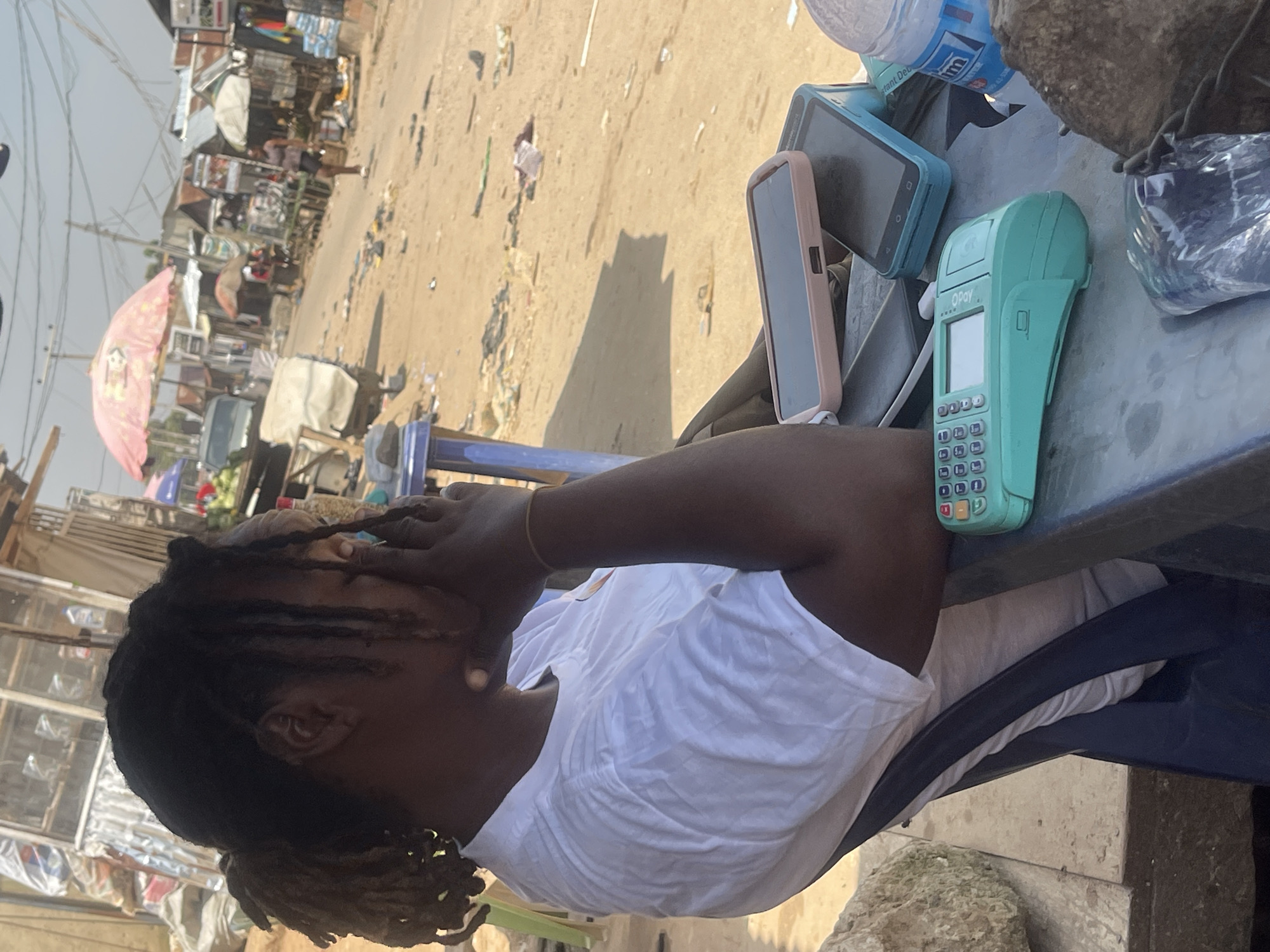 The image depicts an outdoor scene, likely a market, with a person sitting at a table. The person, with dreadlocks and wearing a white t-shirt, has their hands on their face, suggesting they might be stressed or tired. On the table in front of them are several objects, including a teal OPay point-of-sale (POS) terminal, two smartphones in cases (one pink, one blue), and a partially visible plastic bottle of water. The background shows a bustling market environment with stalls, goods, and other people in the distance. The ground appears to be unpaved and dusty, with some litter scattered around. The lighting suggests it is daytime, and the overall atmosphere is that of a typical busy marketplace. The OPay terminal is a recognizable feature, indicating a commercial transaction is likely occurring or has recently occurred.