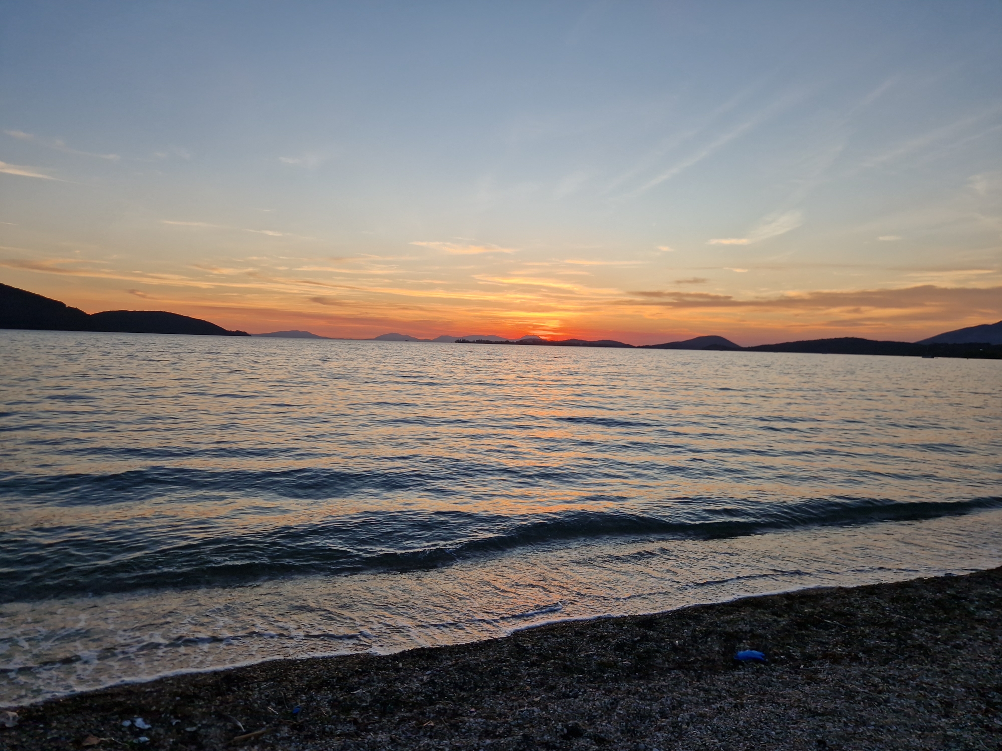 The image depicts a serene seascape at sunset, likely on a beach near the city of Igoumenitsa, Greece, given the silhouetted islands in the distance. The sky transitions from a pale blue at the top to hues of orange and yellow near the horizon, with wispy clouds scattered across. The sun, a bright orange orb, is setting behind a distant island chain, casting a warm glow on the water. Gentle waves lap at a pebble-strewn shore in the foreground. The water is a deep blue with ripples reflecting the colors of the sunset. The overall atmosphere is tranquil and picturesque, characteristic of a Mediterranean evening.