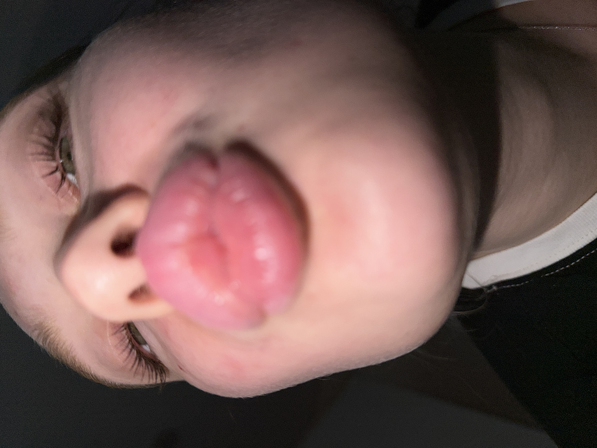 A close-up, tilted downward view of a person's face with their tongue extended towards the camera, partially obscuring the nose. One eye is visible, revealing green irises and eyelashes. The background is dark, with a black garment visible on the right side of the frame with a white collar. The location, Rossiyskiy, Russia, is not discernible from the image.