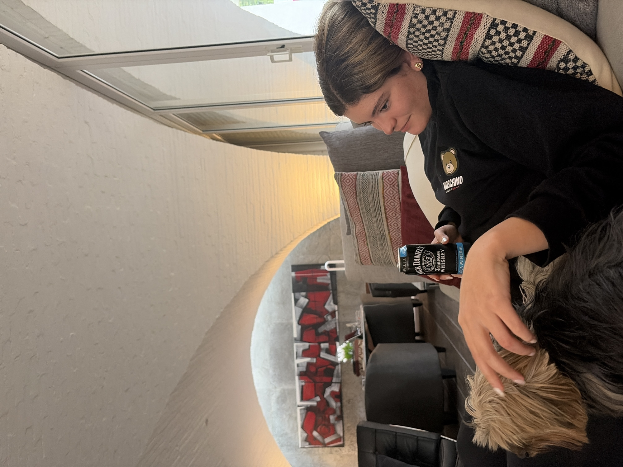 A medium shot captures a woman seated indoors, with a can of "Jack Daniel's" whiskey visible in her hand. She is wearing a black Moschino sweatshirt and appears to be interacting with a dog, whose fur is a mix of blond and black. The interior setting includes a couch with patterned pillows and a rounded, white-walled structure possibly indicating a unique architectural design. The presence of a skylight suggests natural illumination. In the background, there is a contemporary painting with red, white, and gray geometric shapes, along with black chairs and what appears to be a white floral arrangement on a table. The location in the city is listed as Unknown, Unknown.