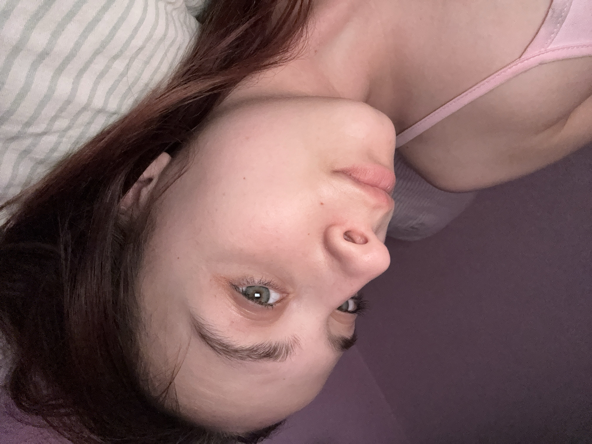 A woman with long brown hair is lying on her back on a striped pillow and purple surface. She is wearing a light pink tank top. The image was likely taken indoors. The location in the city of Прикамский, Russia is speculative and cannot be confirmed from the image.