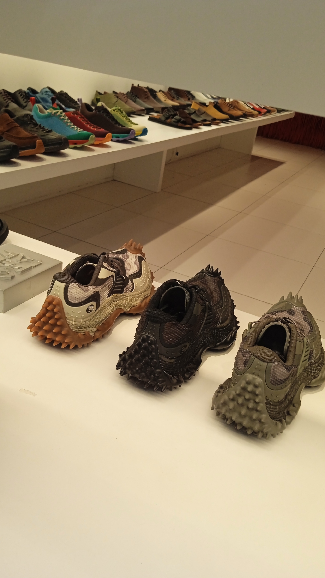 Three uniquely designed, textured shoes are displayed on a white surface in the foreground, with a larger collection of various shoes arranged on a white shelf in the background. The shoes in the foreground feature prominent, rugged soles with numerous spikes or raised bumps, giving them an aggressive, possibly outdoor or athletic aesthetic. The middle shoe is predominantly black, while the shoe on the left has a tan and gray camouflage pattern, and the shoe on the right is a muted green with a similar camouflage pattern. The background shelf holds a diverse assortment of footwear, ranging from sneakers in bright colors like blue, green, and yellow, to more subdued brown and tan loafers and sandals. The overall environment appears to be a retail or exhibition space, possibly a shoe store or a design exhibition, with a plain, neutral-colored wall and floor contributing to a clean, modern presentation. The lighting is bright and even, highlighting the products. While the prompt specifies Milan, Italy, there are no visual cues within the image to confirm this location; it is presented as a matter of fact.