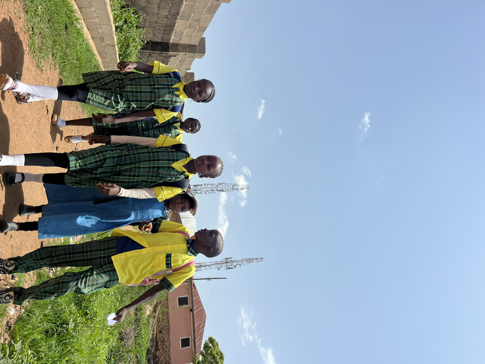 In this outdoor, upside-down image, a group of seven children, likely students, are standing on a dirt path next to a grassy area and a brick wall. The children are dressed in uniforms. The child on the far left is wearing a yellow shirt with a green and black plaid collar and cuffs, and matching plaid pants. They are holding a small white object. The second child from the left is wearing a blue dress. The remaining five children are all wearing green and black plaid shirts with yellow collars and cuffs, and black pants. Three of these children are also wearing plaid skirts over their pants. Behind the children, there is a red-roofed building with two windows, and several tall cellular towers are visible against a bright blue sky with scattered clouds. The image is oriented such that the sky is at the bottom and the ground is at the top.