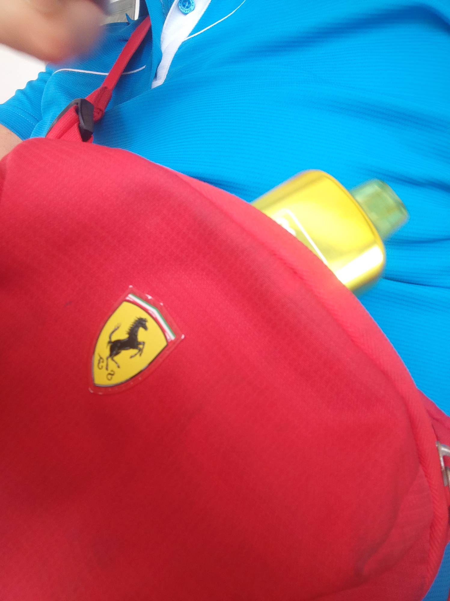 The image shows a close-up of a person wearing a blue shirt with white accents. A red bag is slung over their shoulder, with a Ferrari logo visible on it. A yellow bottle is also partially visible, resting on the bag. The scene appears to be outdoors, with a bright, sunny environment. The specific location in the city of Bar, Montenegro is not discernible from the provided image.