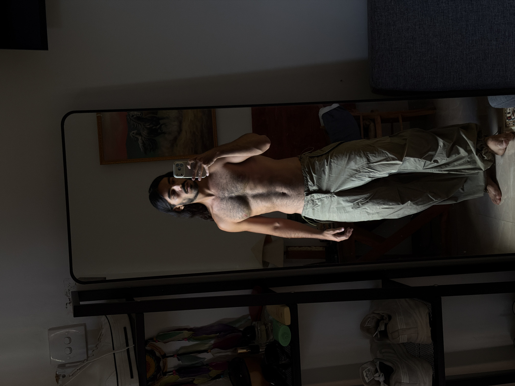 A shirtless man with long dark hair, wearing loose olive green pants, stands in front of a full-length mirror, holding a cell phone to take a selfie. The room is dimly lit. In the background, there's a painting on the wall and a dark gray couch. The mirror is framed in black metal and has shelves beneath it holding shoes and other items. An electrical outlet is visible on the wall next to the mirror. The image was taken in North Kuta, Indonesia.