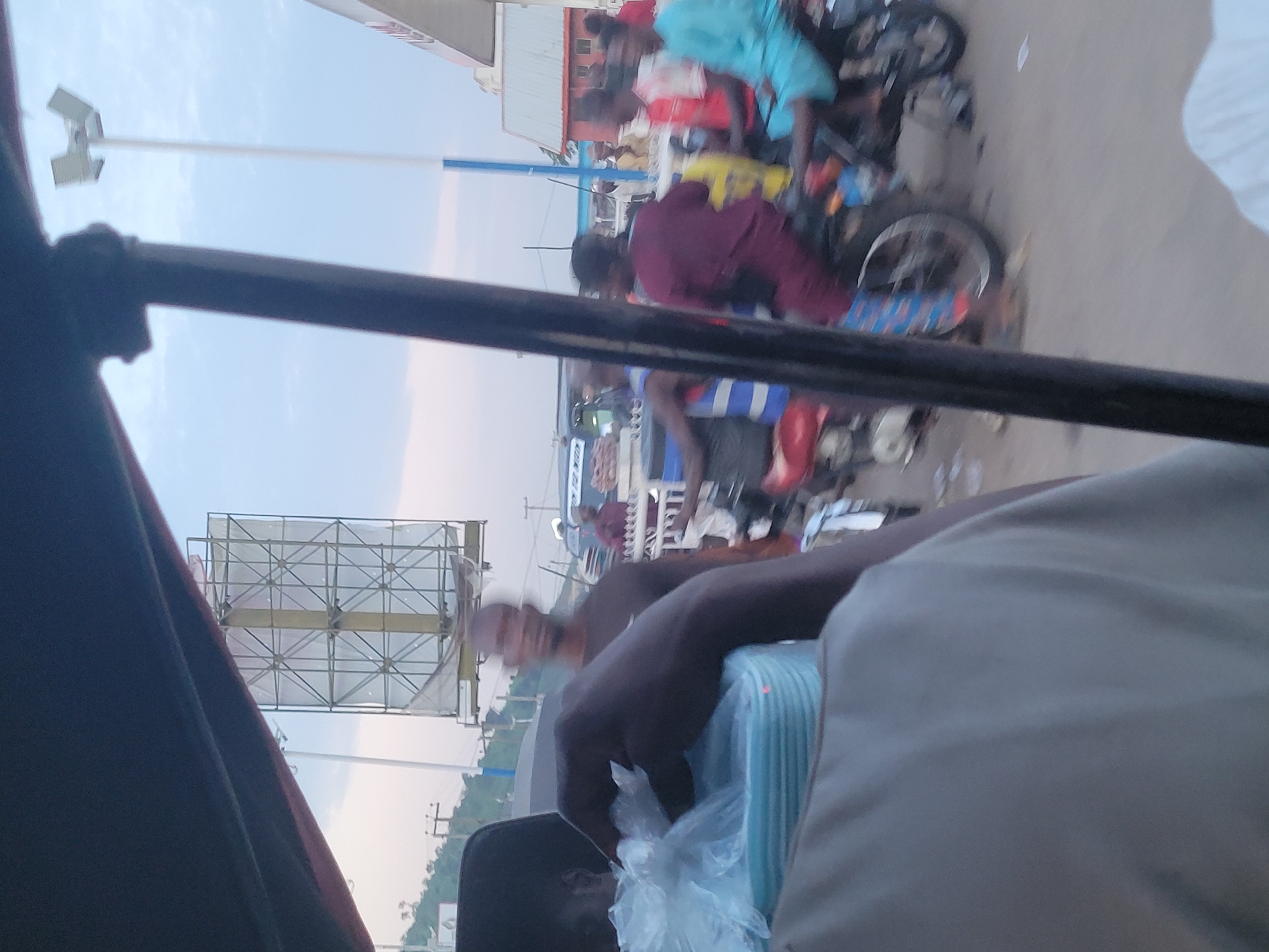 The image captures a bustling street scene in Lokoja, Nigeria. In the foreground, a person's hand is seen holding a stack of blue plastic containers wrapped in clear plastic. This suggests a transaction or delivery of goods. The background reveals a busy roadside environment with several people and motorcycles. A billboard structure is visible in the distance, along with a tall light pole. Buildings and trees line the street, indicating an urban or semi-urban setting. The overall atmosphere appears dynamic and lively, typical of a commercial area in Nigeria.
