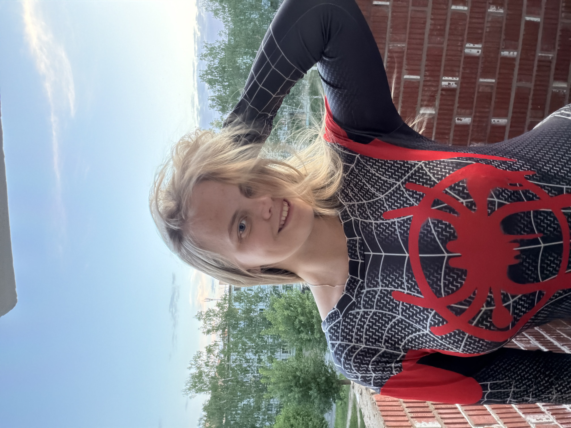 A woman is posing for a photo on a balcony. She is wearing a form-fitting, black and white suit with a large red spider emblem on the torso. The suit has a spider web pattern. She is fair-skinned, with blonde hair, and is smiling. Behind her is a brick wall and in the background, there are trees and a building. The sky is partially visible, showing blue sky and clouds. Based on the architecture and environment, the location is presumed to be in Izhevsk, Russia.