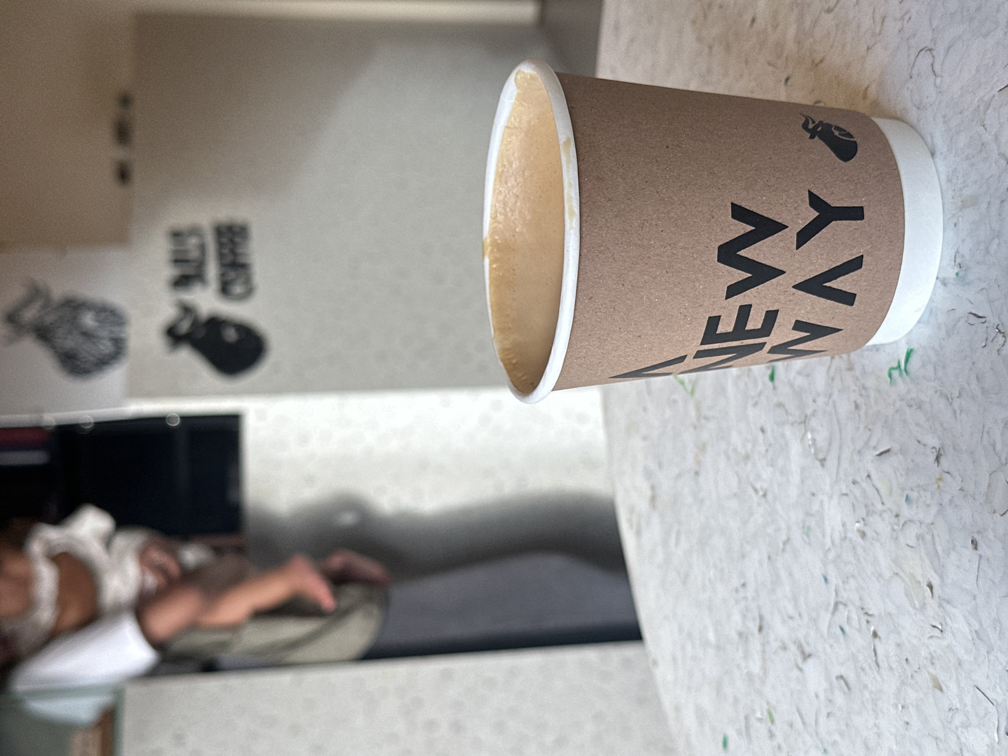 A coffee cup with the words "NEW WAY" printed vertically in black letters on a brown paper sleeve is on a light-colored, possibly plastic surface speckled with tiny pieces of color. The cup is partially filled with coffee, visible at the rim. In the background, out of focus, is a room interior. There is a white wall with a dark image of a buffalo and writing on it. There is a TV, a person lying on the couch with feet visible and white shirt. Based on the location "North Kuta, Indonesia," it is likely the "New Way" Cafe located in Canggu.