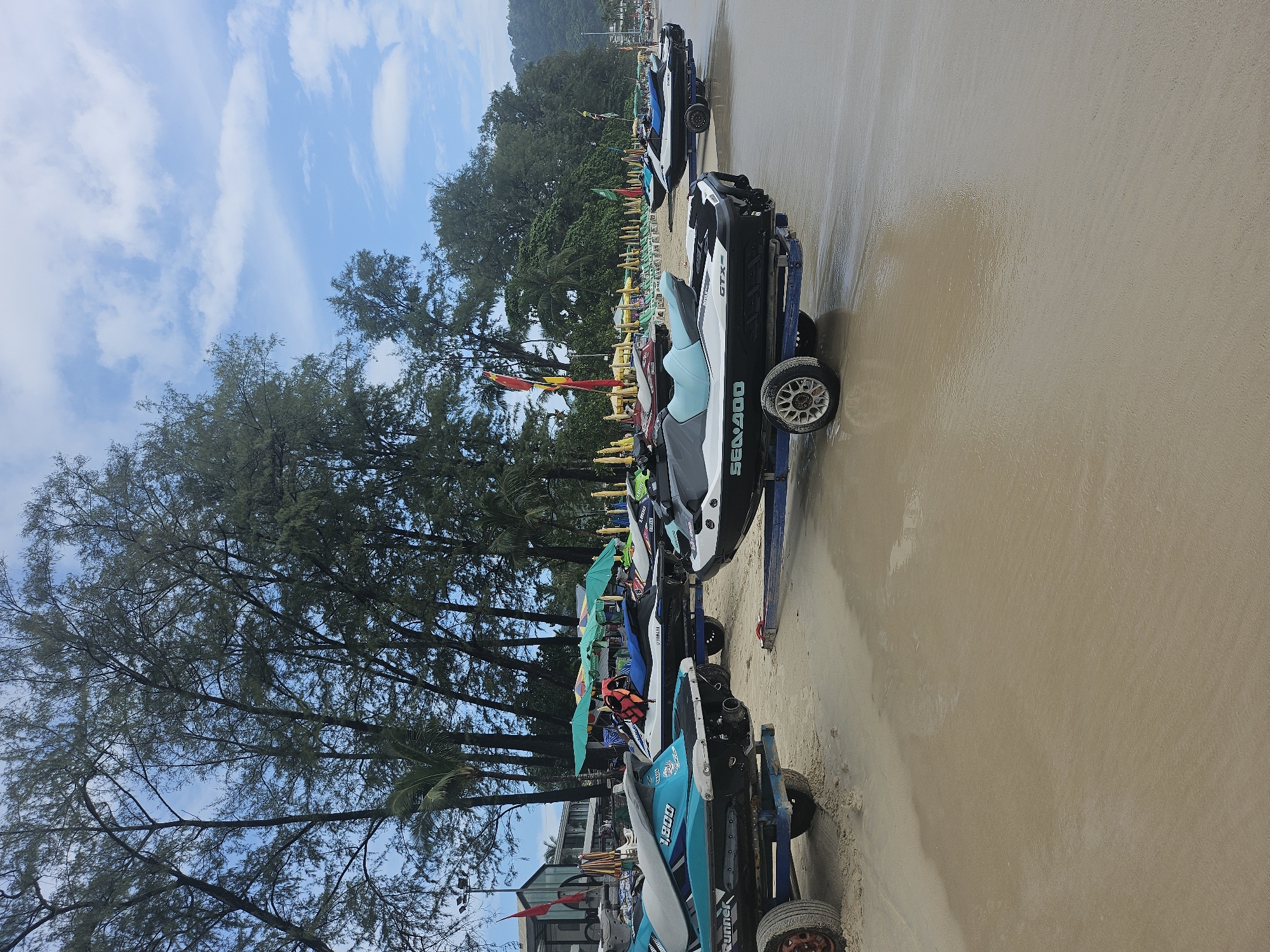 The image depicts a beach scene in Patong, Thailand, with multiple jet skis parked on trailers on the wet sand near the water's edge. The jet skis are of various colors, including teal, white, black, and green. Some have "Sea-Doo" and "GTX" branding visible. In the background, there is a line of green umbrellas, lounge chairs, and trees, with buildings and a hillside visible further in the distance. The sky is partly cloudy with a bright blue hue. The overall impression is one of a recreational area on a beach.