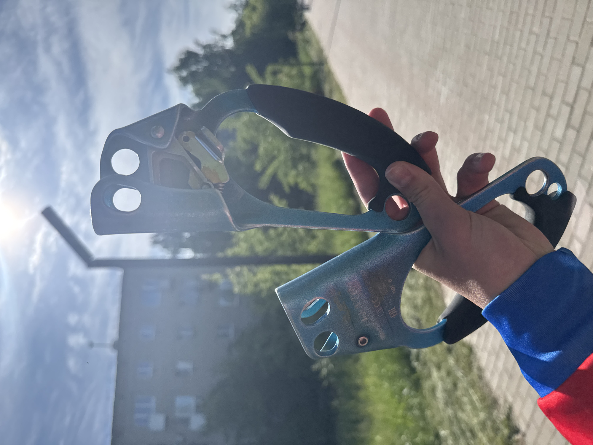 A person's hand, wearing a blue and red sleeve, holds two light blue and black climbing ascenders upwards against a bright outdoor background in Chaikovsky, Russia. The ascenders, which appear to be a pair designed for left and right hands, are held parallel to each other. Bright sunlight from the left creates a lens flare and illuminates the scene, indicating daytime on a clear, sunny day with some clouds visible in the sky.
In the blurred background, there are green trees and foliage, a light-colored paved path or sidewalk, and a multi-story building with dark windows, suggesting an urban or suburban setting. A dark, vertical pole or structure is also visible against the sky and building.
Visible text on the lower ascender clearly shows "PETZL" (brand), "ASCENSION" (model name), "EN 12841 B" (a European standard for rope access equipment), and "CE 1019" (CE certification marking). The image captures a moment possibly before or after a climbing activity, showcasing the equipment.