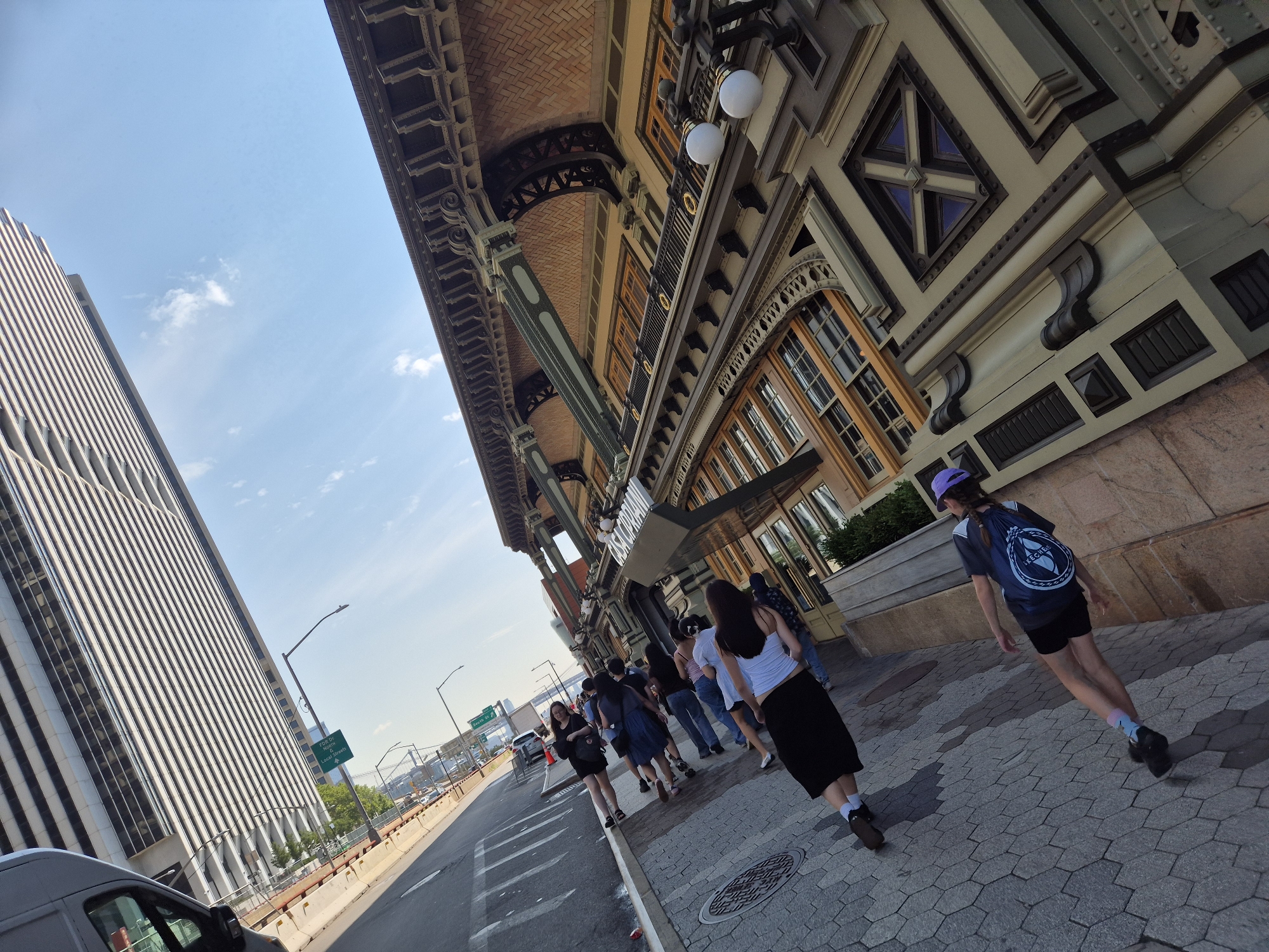 The image depicts a street scene in New York City, likely near the World Trade Center. On the left, a tall, modern skyscraper with a ribbed facade dominates the background. To the right, a building with a more ornate, older architectural style, featuring a brick facade under a decorative overhang, lines the street. A group of people are walking along the sidewalk in front of this building. One person in the foreground wears a black skirt and a white top, carrying a black backpack. Another person ahead of them, also in the foreground, wears a purple hat and a dark blue t-shirt with a circular emblem on the back, carrying a blue backpack. The sidewalk is paved with hexagonal stones, and a manhole cover is visible. Across the street, a highway barrier separates the sidewalk from a road with vehicles. Traffic signs are visible, one indicating "FORUM NORTH LOCAL STREET." The sky is clear with a few wispy clouds. The overall scene suggests a sunny day in a bustling urban environment.