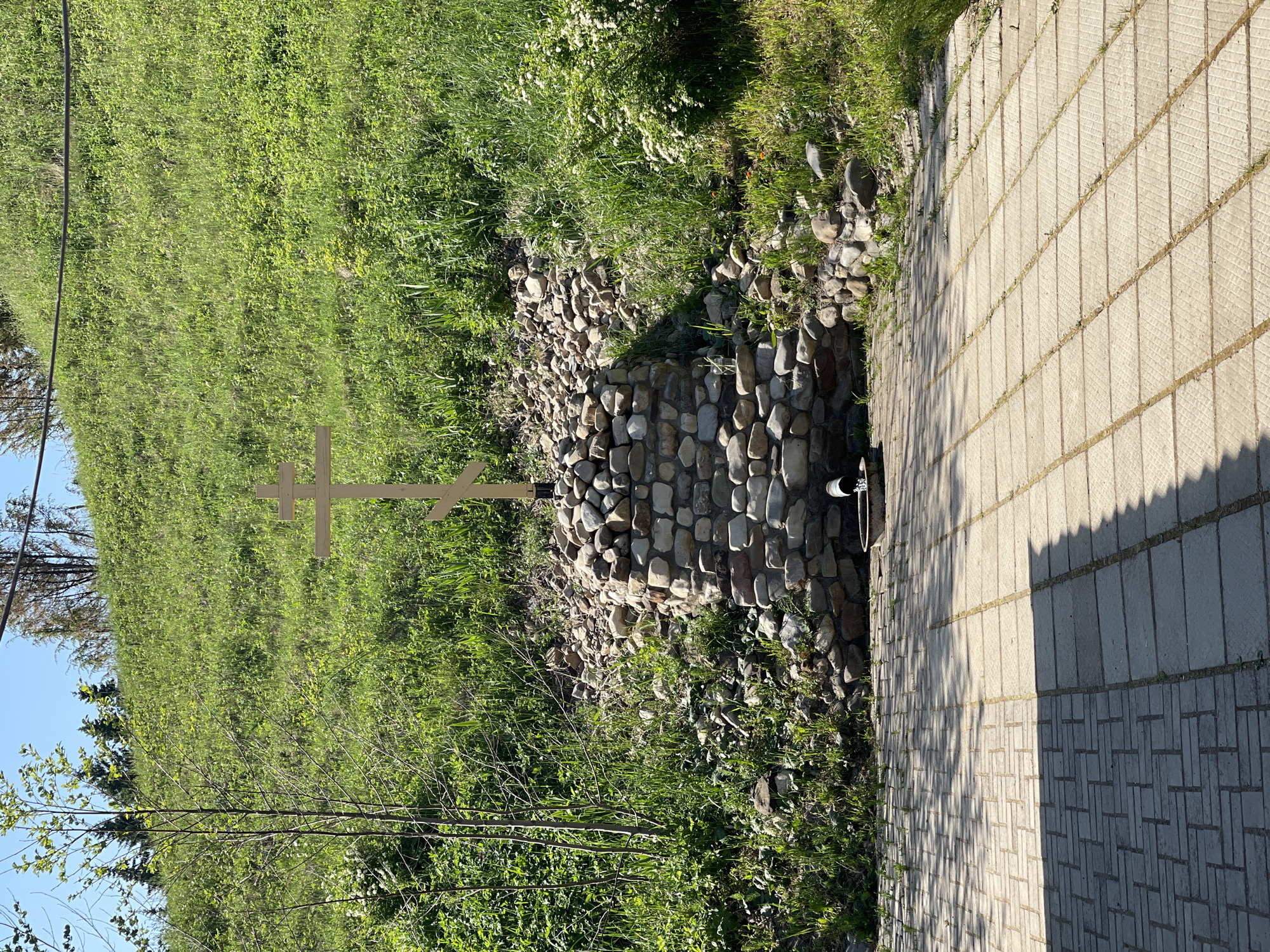 A ground-level, outdoor shot in Перевозное, Russia shows a stone-lined spring or water source, with a wooden Christian cross nearby. The spring is constructed of large, smooth stones and has a pipe protruding from its opening. The cross is simple and made of unfinished wood. The area around the spring is covered with green grass, foliage, and small trees. Adjacent to the spring is a paved walkway or patio made of light-colored rectangular stones. A shadow falls across the walkway, suggesting the presence of a nearby object or structure. The sky is visible in the distance.