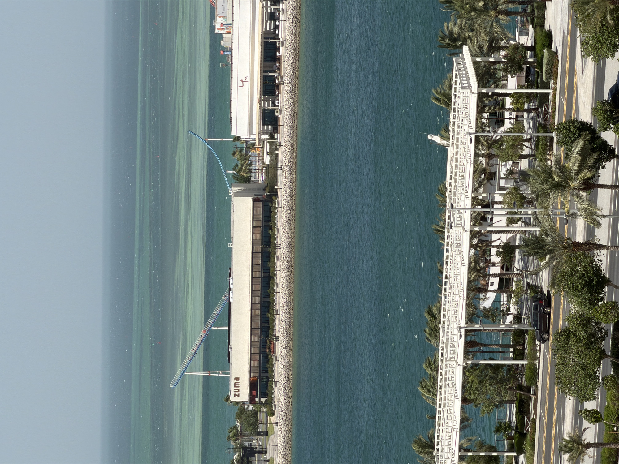 An elevated, wide-angle, eye-level shot captures a waterfront scene under a hazy, pale blue sky. In the foreground, a dark gray SUV is parked on the right side of a road bordering a pedestrian walkway and a white, open-air canopy structure supported by pillars. Beyond this, a body of deep blue water stretches towards a lighter, turquoise-colored sea in the distance, marked by subtle lines suggesting shallow areas or sandbars. A red buoy is visible in the distant water.
Further back, on the edge of the water, a long, beige building with a dark-trimmed facade and numerous windows is situated. The word "zuma" in dark lettering is displayed prominently on this building's exterior. Adjacent to it, another white building with a red-trimmed roofline and outdoor seating area is visible. Connecting these structures and extending over the water is a light blue roller coaster track supported by white pillars. Palm trees and other greenery are scattered along the waterfront. The overall impression is one of a modern, resort-like environment.