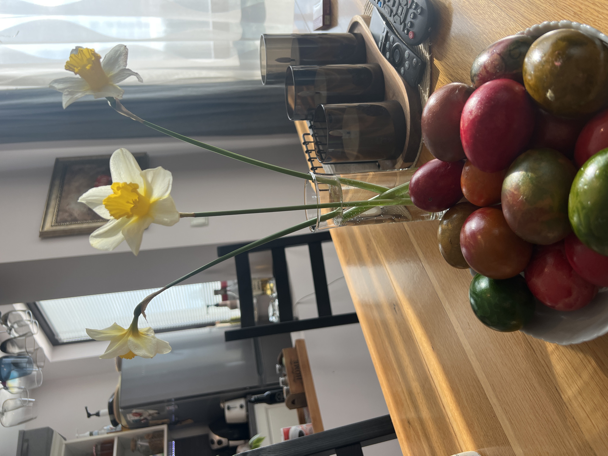 A still life on a wooden table, including daffodils in a glass vase, a bowl of painted eggs, and a TV remote, inside a home, possibly in Sofia, Bulgaria, based on architectural styles.
The focal point is a clear glass vase holding three daffodils with white petals and yellow centers. The stems of the daffodils are visible within the water. To the right of the vase is a small bowl filled with painted Easter eggs in various colors including red, green, brown, and orange. Beside the vase is a wooden tray containing dark-colored drinking glasses.
A black TV remote rests on the table near the tray.
Background details include a white window, a framed picture on the wall, and a shelving unit with bottles and decorative items. A TV is visible below the window, indicating a living area. The environment is well-lit, likely by natural light.