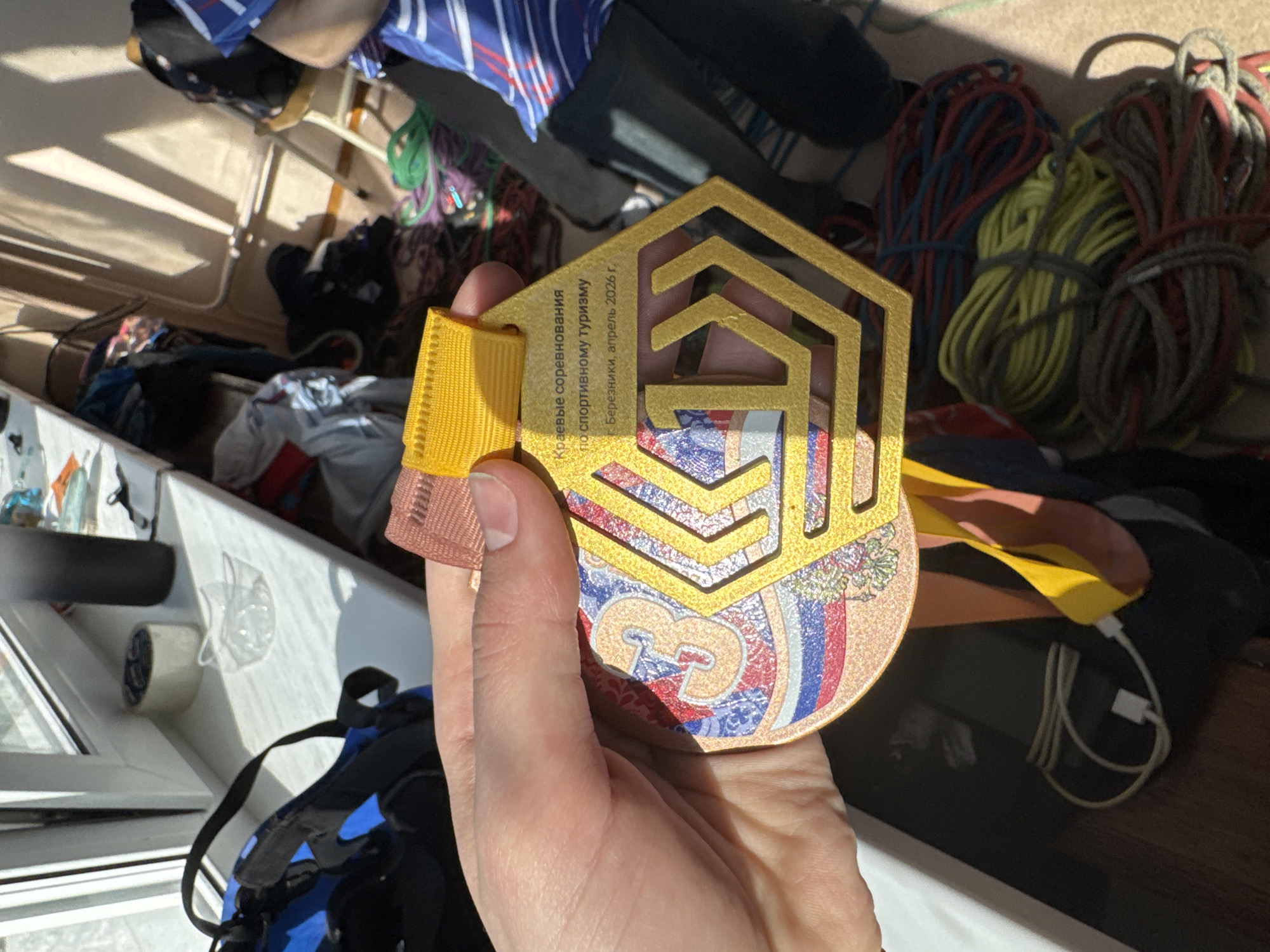 A hand holds a medal with a hexagonal gold frame and a ribbon in yellow, bronze, and pink. The medal has an image that includes the colors of the Russian flag and the text "Краевые соревнования по спортивному туризму, Березники, апрель 2026 г." The background features ropes, clothes, a window, and a backpack. The scene appears to be indoors. Usolye, Russia, is not directly discernible from the image.