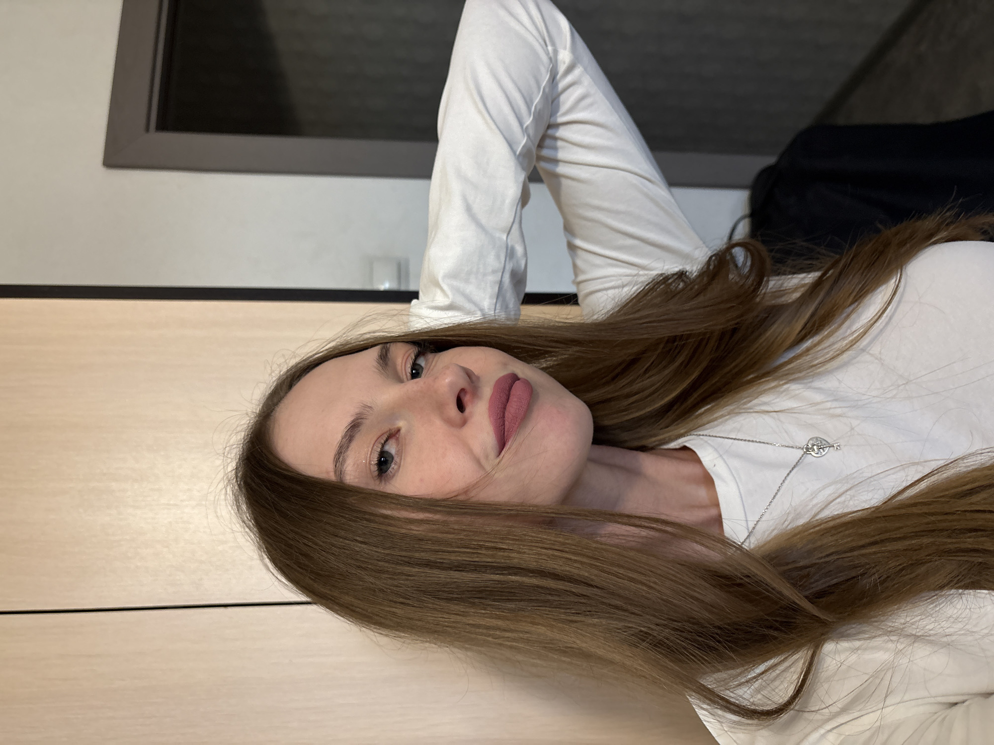 The image shows a young woman with long, light brown hair, wearing a white long-sleeved shirt and a silver necklace with a key-shaped pendant. She is looking towards the camera with a slight smile and has makeup on, including pink lipstick and glitter on her eyelids. Her right arm is raised, with her hand behind her head. The background appears to be indoors, with a wooden closet or cabinet to her left and a doorway to her right. The lighting suggests an indoor setting, possibly a home or apartment. There is no indication of the city of Perm, Russia, within the image itself.