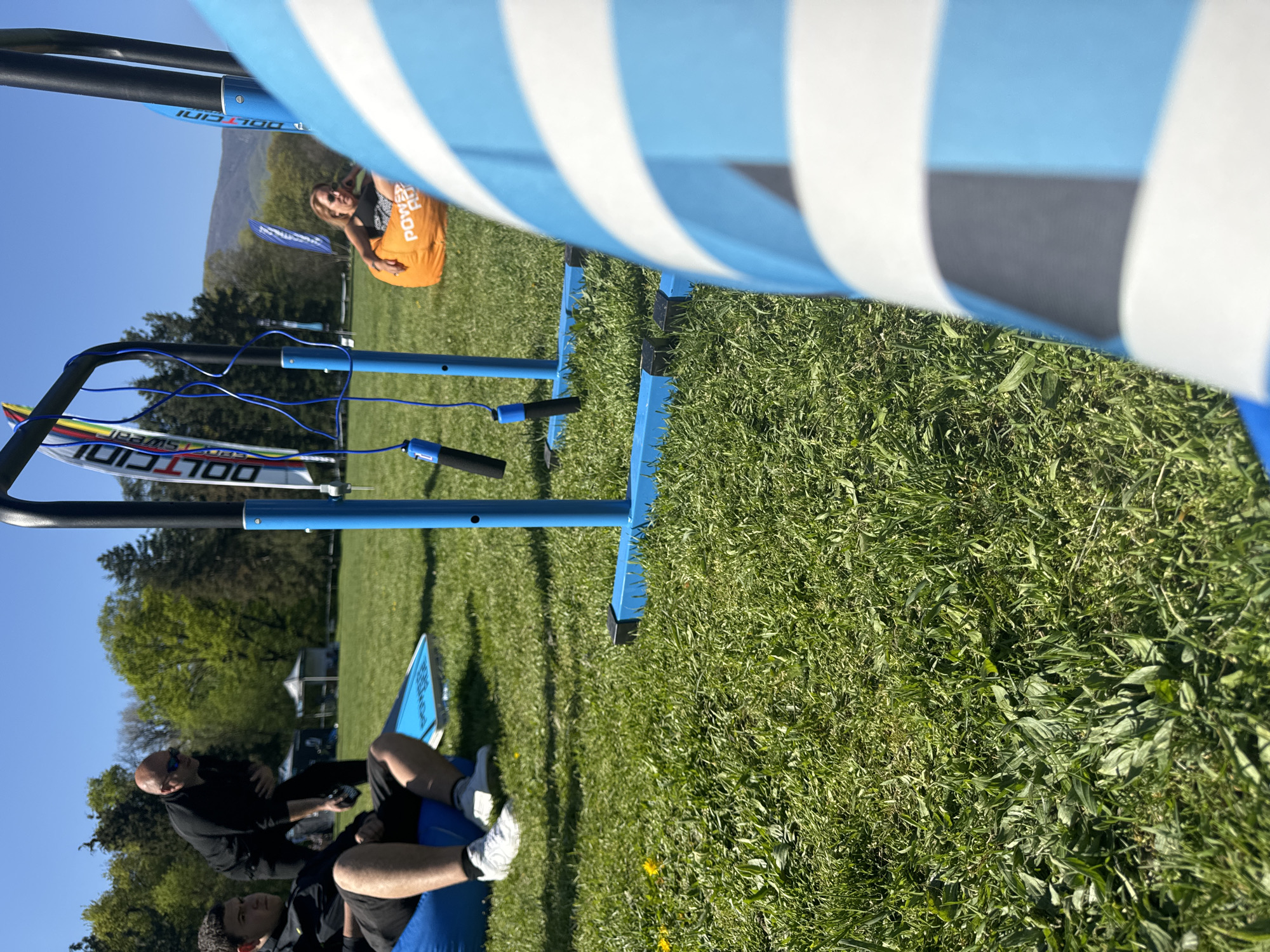 The image shows an outdoor scene on a grassy field, potentially a park or recreational area. The view is partially obstructed by a blue and white striped object, which appears to be part of an event setup. In the background, there are trees and a hill or mountain. There are several people in the scene. One woman in the background is wearing an orange shirt. Closer to the camera, there are two men sitting on a blue object, possibly inflatable or a piece of event equipment. One of the men is wearing a black shirt and sunglasses. The location may be somewhere in Sofia, Bulgaria, however, nothing can be determined about the specific location.