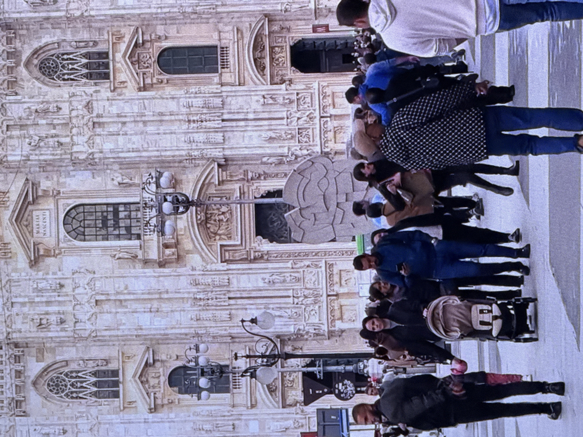 The image depicts a busy street scene in front of a large, ornate building that appears to be a cathedral. Several people are walking in various directions, some in groups and others individually. One woman is pushing a baby stroller. The architectural style of the building suggests it is a European landmark, likely gothic in design, with intricate carvings and multiple windows. There are streetlights visible, and the ground is paved. The overall atmosphere is one of a public gathering place or tourist attraction.
However, the question states the location is Dubai, United Arab Emirates. Dubai is known for its modern architecture and skyscrapers, which do not align with the gothic style of the building shown in the image. It is highly unlikely that this image was taken in Dubai. The building's architecture is characteristic of European cathedrals, such as the Milan Cathedral in Italy.