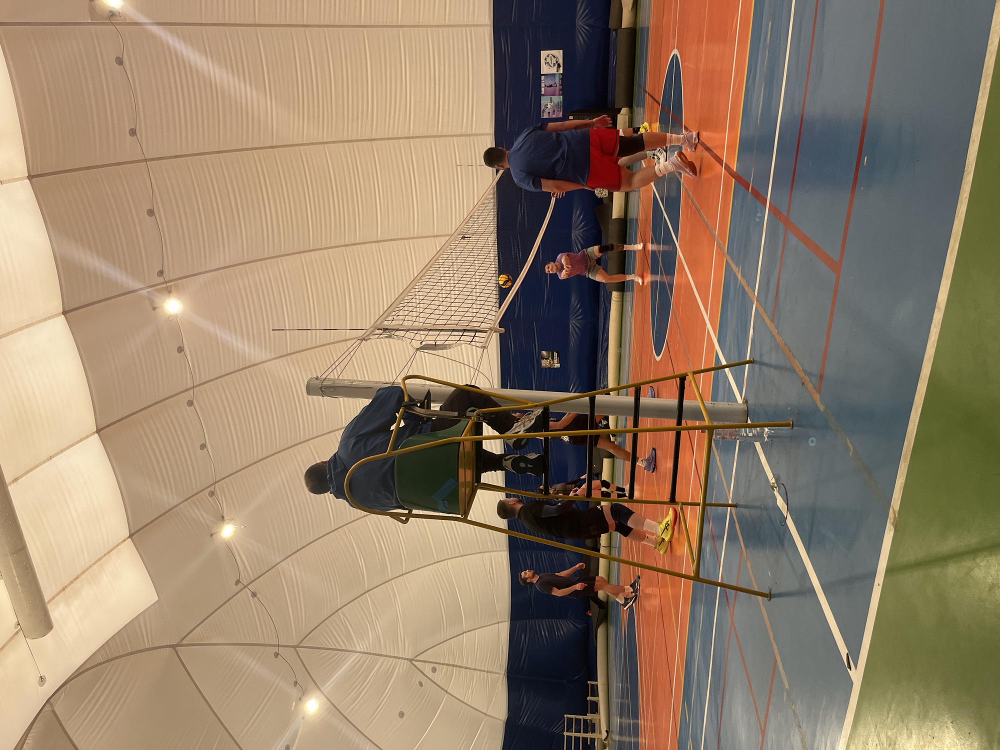 The image shows an indoor volleyball court with a high, domed ceiling. The court has blue and orange sections, delineated by white lines. A volleyball net stretches across the court. Several people are visible: two near the net, one appearing to serve or hit a volleyball, and another standing nearby. Closer to the camera, a man sits in a high chair, possibly officiating the game. A few other individuals are scattered around the court. The environment appears to be a gymnasium or sports hall. The location is potentially within the city of Sofia, Bulgaria, but specific landmarks or identifiers are not visible in the image to confirm this.