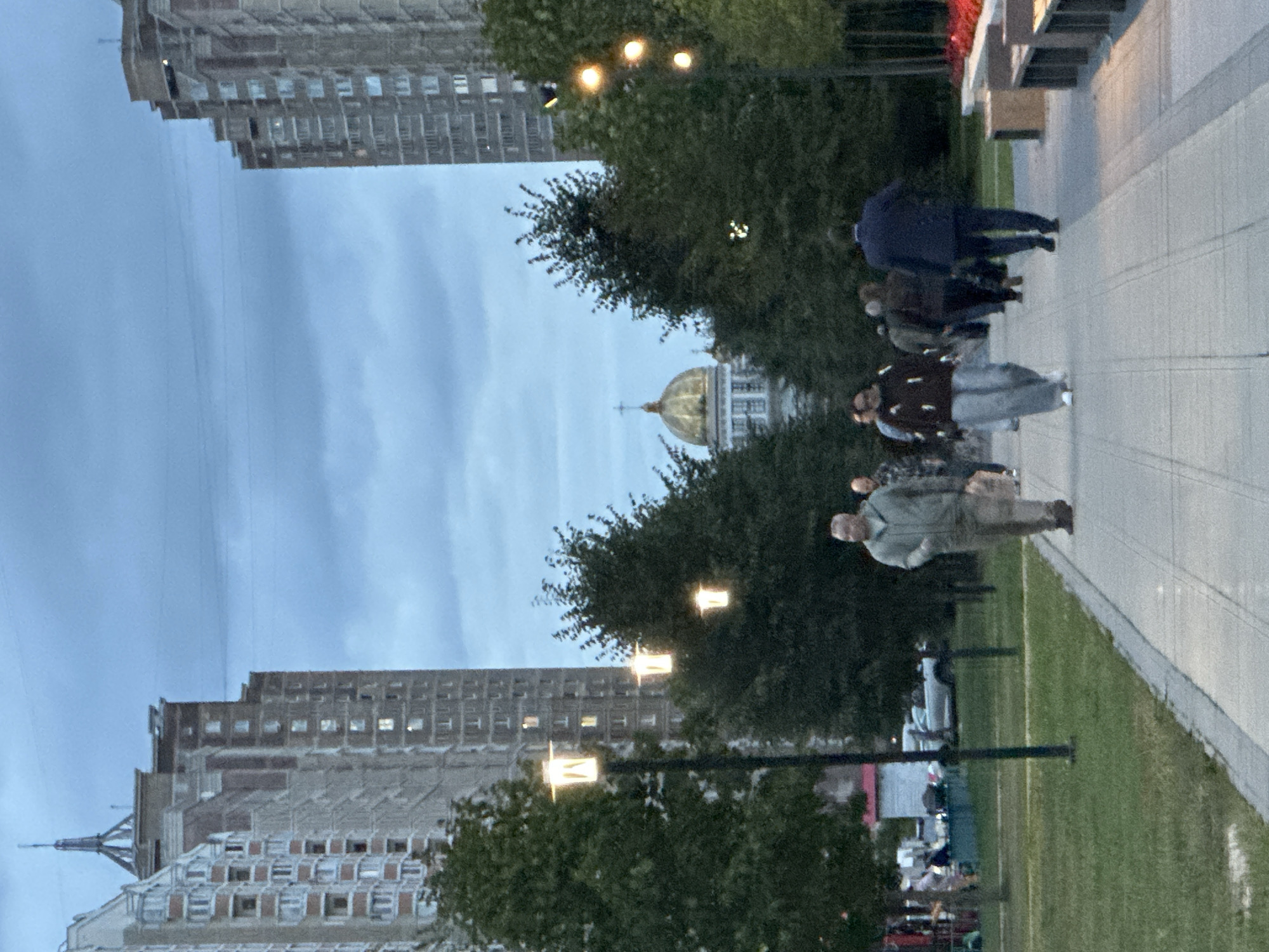 In Podolsk, Russia, on a cloudy evening, people walk along a paved pathway in a park-like setting. The path is flanked by a grassy area on the left and trees on the right. Tall, modern apartment buildings with numerous windows are visible on either side of the scene, creating a canyon effect. In the distance, a building with a prominent golden dome, possibly a church or cultural center, is situated between the apartment buildings. Several streetlights with glowing yellow lamps are positioned along the pathway, illuminating the area. A group of people, some walking individually and others in small groups, are seen moving in both directions on the path. In the background, more trees and some low-lying structures are present. The overall atmosphere is one of urban life continuing as daylight fades.