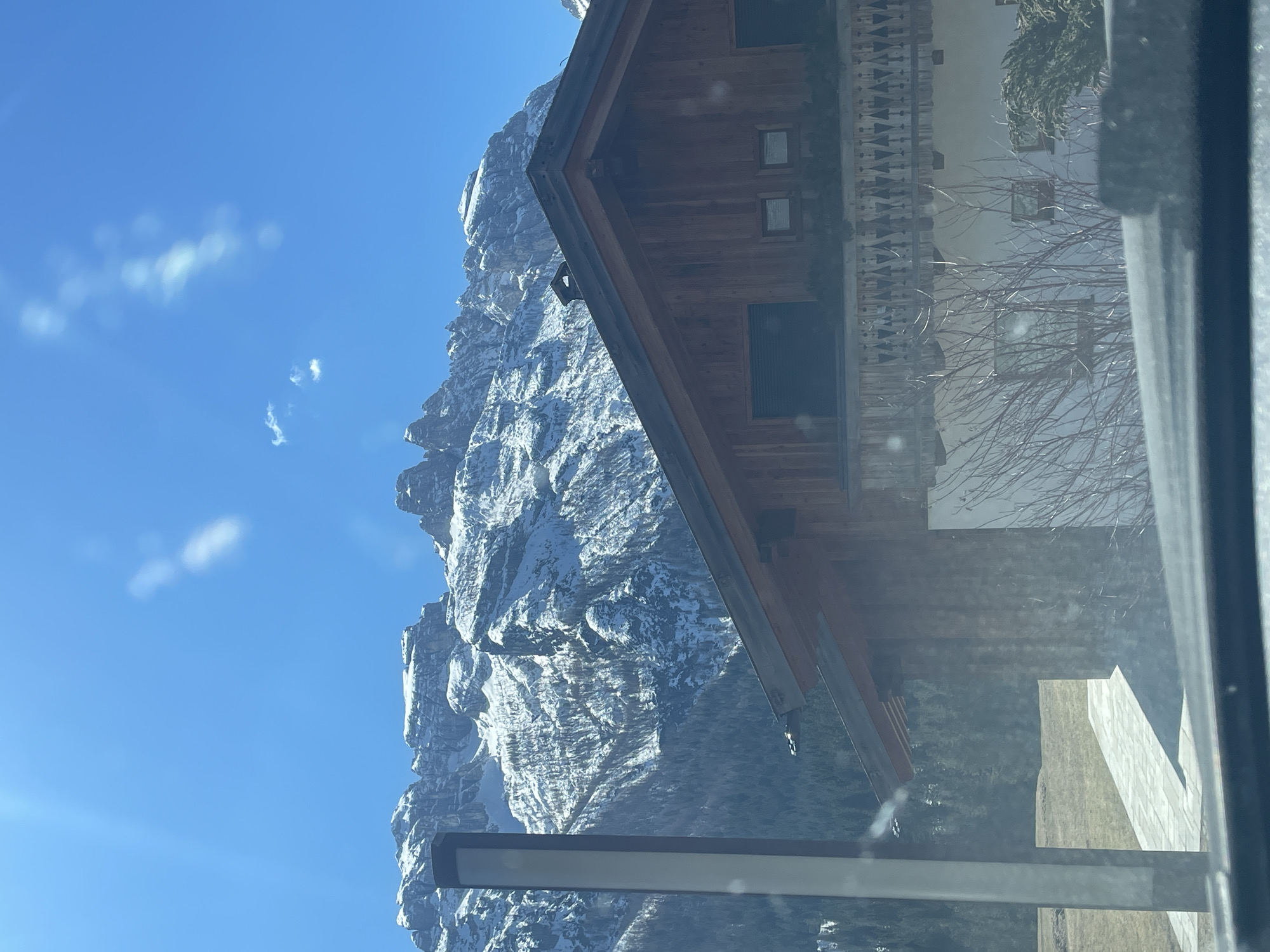 A landscape photograph depicts a snow-dusted mountain range against a bright blue sky with sparse clouds. The image is taken from within an interior space, indicated by the presence of building structures including a chalet-style building on the right and a metal window frame along the bottom edge. The perspective is tilted. The location is suggested as being in Sappada, Italy based on the prompt. There is no visible human activity, people, subjects, or interactions.