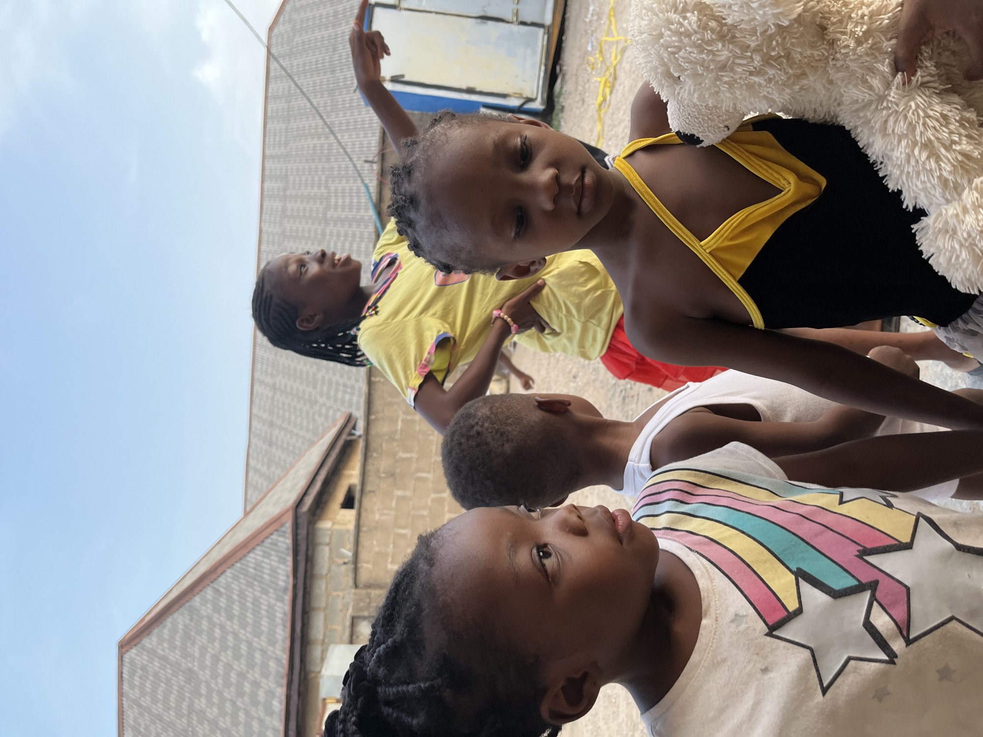 A group of four children are standing outdoors in Iddah, Nigeria. The background includes buildings constructed of gray blocks and a clear, light blue sky. The child in the foreground is wearing a white shirt with a colorful rainbow and star design. Behind this child is another wearing a plain white tank top. A third child is wearing a yellow shirt and has their arm raised, while the fourth child is in a black and yellow swimsuit and holding a white, fluffy stuffed animal.