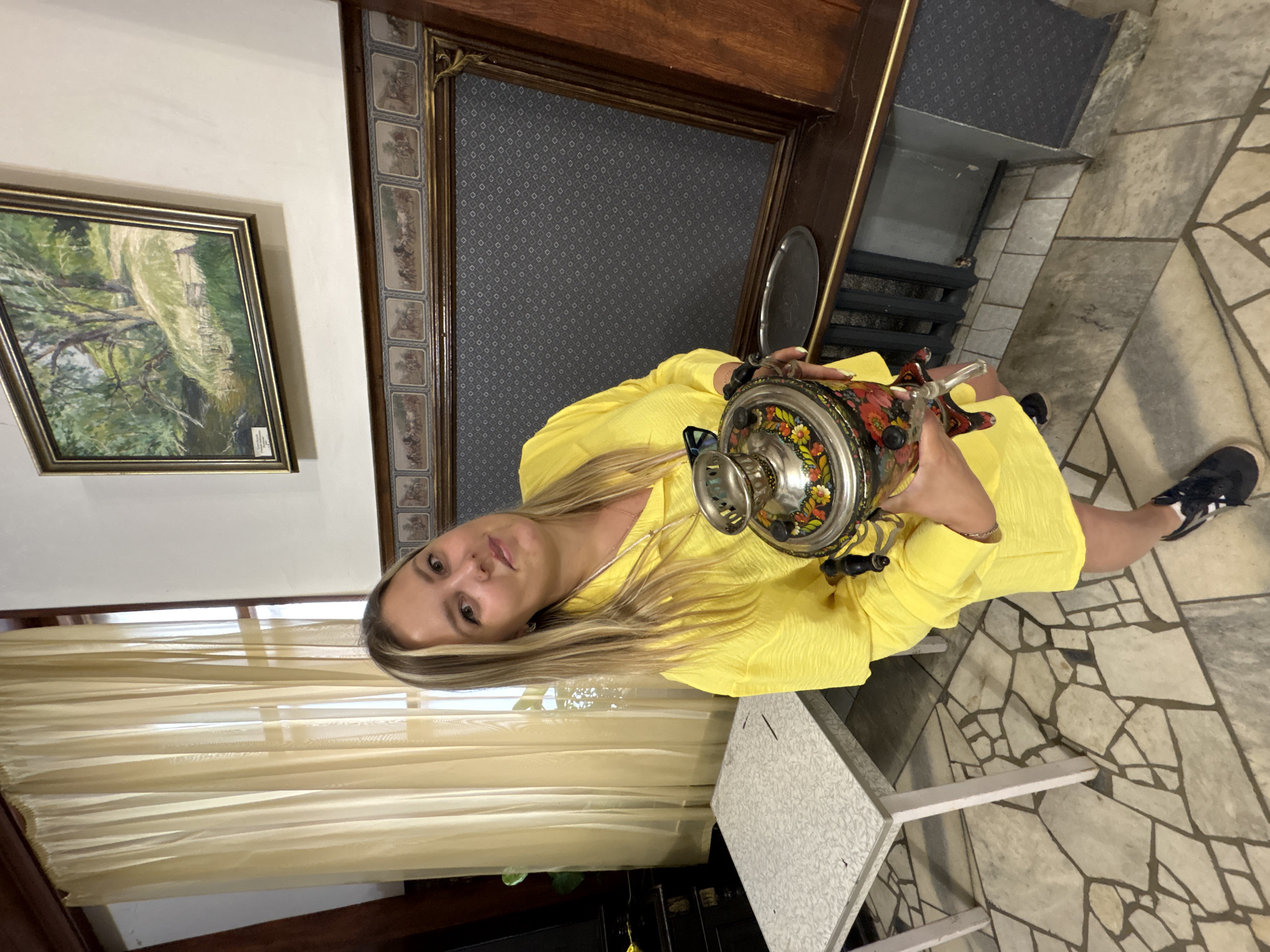 A woman wearing a bright yellow dress holds a decorative samovar, a traditional Russian tea urn, in her left hand. She is positioned in what appears to be a room with a light-colored, mosaic-tiled floor. To her left, a framed painting of a landscape hangs on a cream-colored wall. Behind her, a dark wooden mantelpiece with ornate detailing and a patterned dark grey fabric panel is visible. The setting suggests an indoor environment, possibly a home or a museum, with elements of traditional Russian decor. The lighting appears to be natural, possibly from a window behind the viewer, illuminating the scene. The woman's expression is neutral. No specific time of day or weather conditions are discernible. There is no visible text in the image.