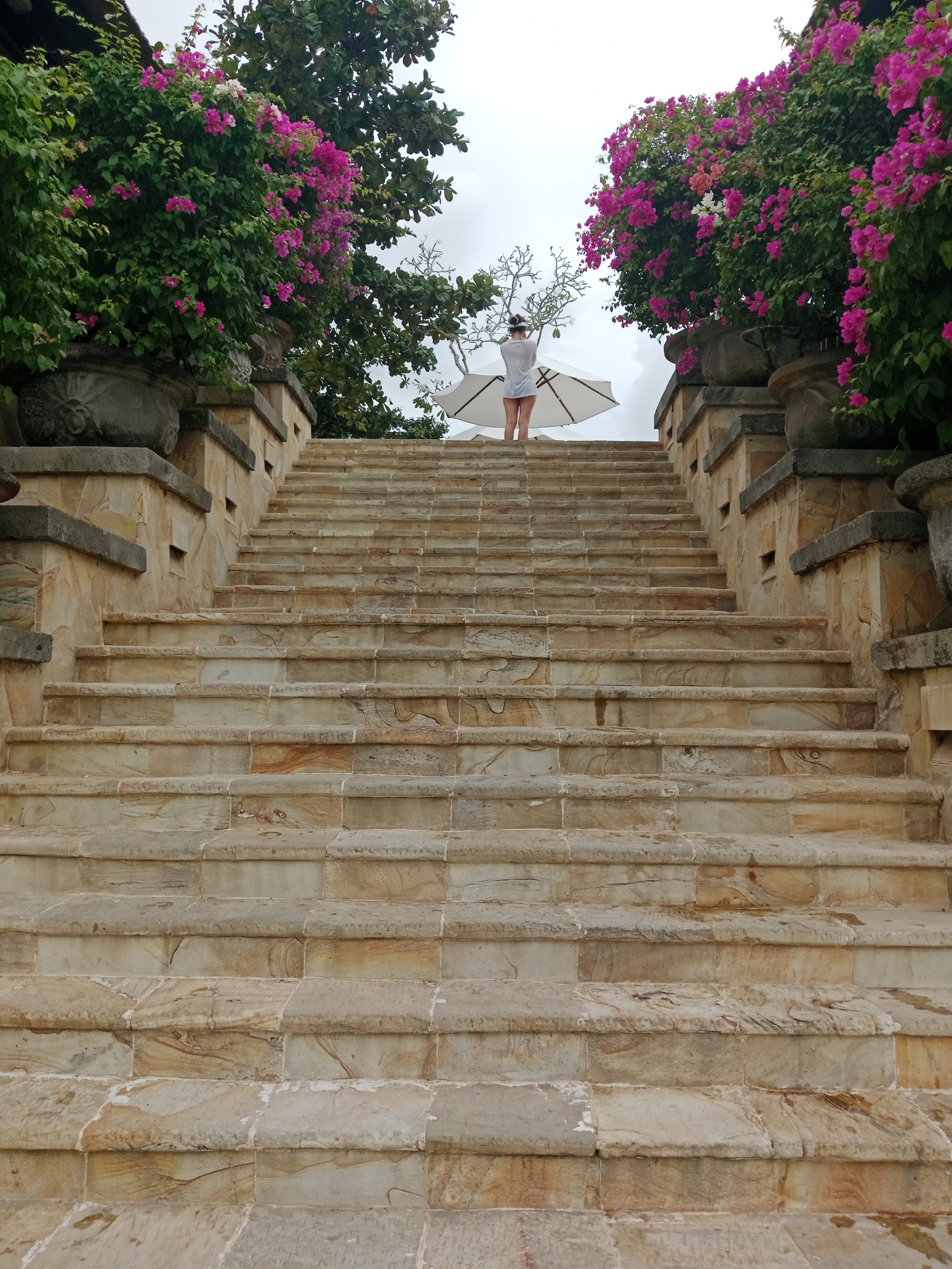 A person stands at the top of a wide, stone staircase, holding an open white umbrella. The staircase is flanked by large stone planters filled with vibrant pink bougainvillea flowers and lush green foliage. The architecture suggests a tropical resort or villa, consistent with locations in Kuta Selatan, Indonesia. The sky is overcast, indicating a cloudy day. The person, viewed from behind, wears a white top and shorts. The scene is tranquil and suggests a moment of respite or enjoyment of the surroundings.