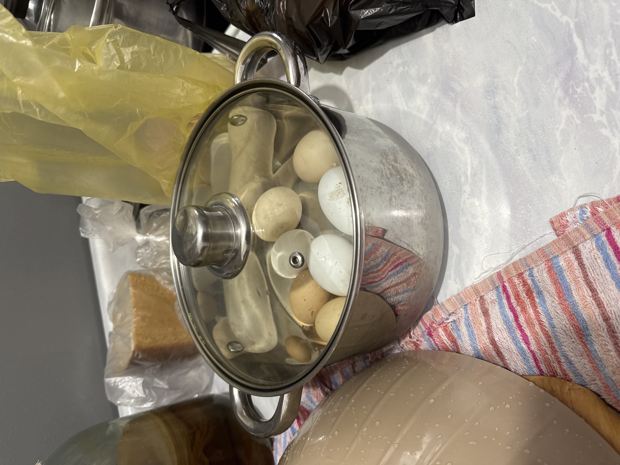 A close-up, eye-level shot shows a stainless steel pot with a glass lid filled with several eggs, some white and some brown, submerged in water. The pot is situated on a white countertop, partially covered by a colorful striped dishtowel. To the left of the pot is a large, light-colored ceramic bowl with water droplets on its surface. In the background, various items are visible, including a large glass jar with a brown liquid, a block of what appears to be sugar or brown rice wrapped in plastic, and a bright yellow plastic bag. A black plastic bag is visible to the right of the pot. The overall setting appears to be a domestic kitchen. The image does not contain any elements that would indicate a specific location in Trudovoye, Russia.