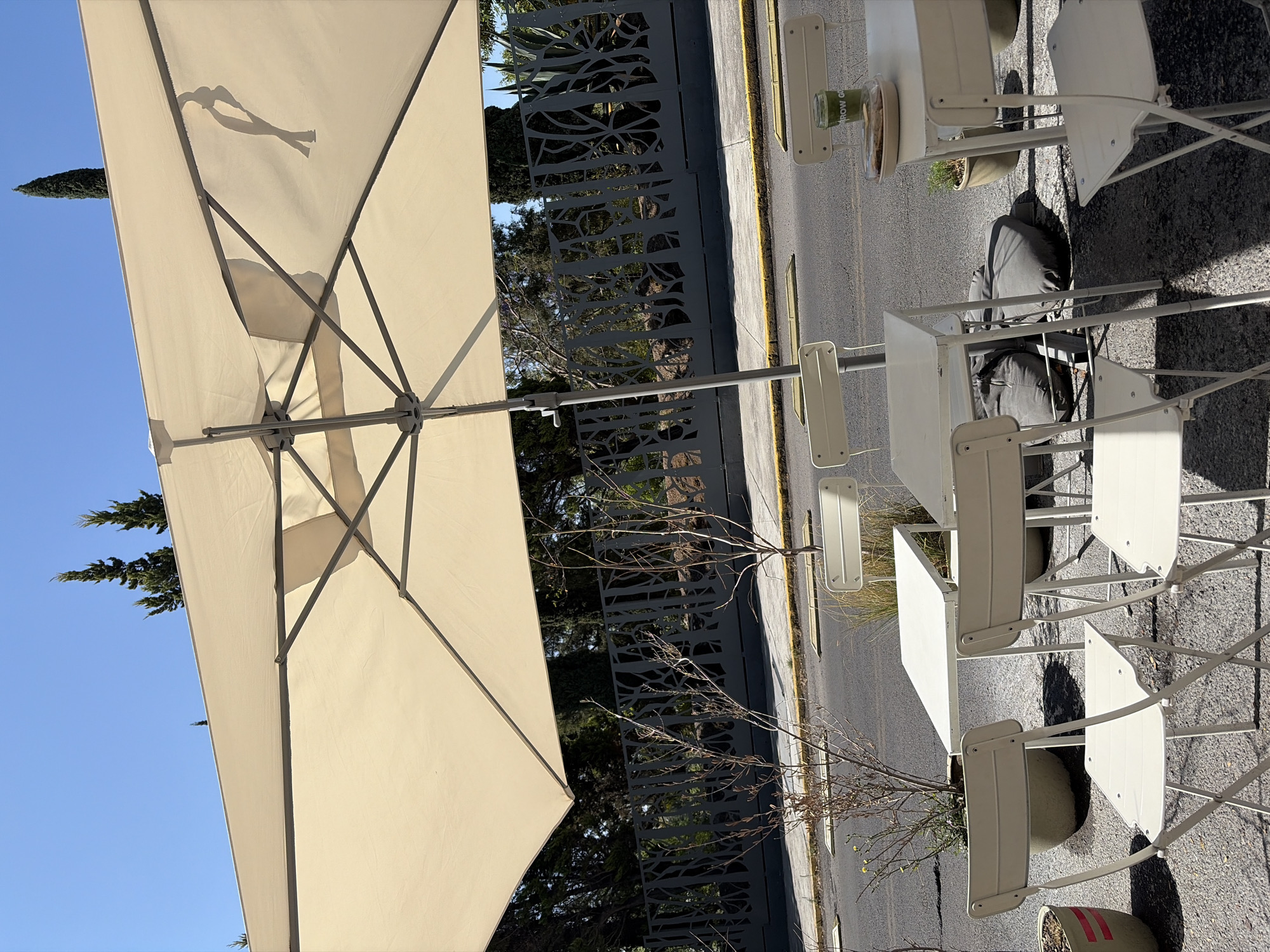 The image depicts an outdoor cafe setting, likely in Puebla, Mexico. A large, beige parasol dominates the left side of the frame, its metal frame clearly visible against a blue sky. A stylized logo of a human figure is imprinted on the parasol's fabric. To the right, a row of white metal tables and chairs are arranged on a paved surface, likely a sidewalk or patio. Some of the chairs are occupied by what appears to be discarded bags or belongings. Potted plants are interspersed among the tables. A decorative metal fence runs along the edge of the paved area, with trees and foliage visible behind it. The scene is bathed in sunlight, creating strong shadows. The overall impression is that of a quiet, possibly deserted cafe scene.