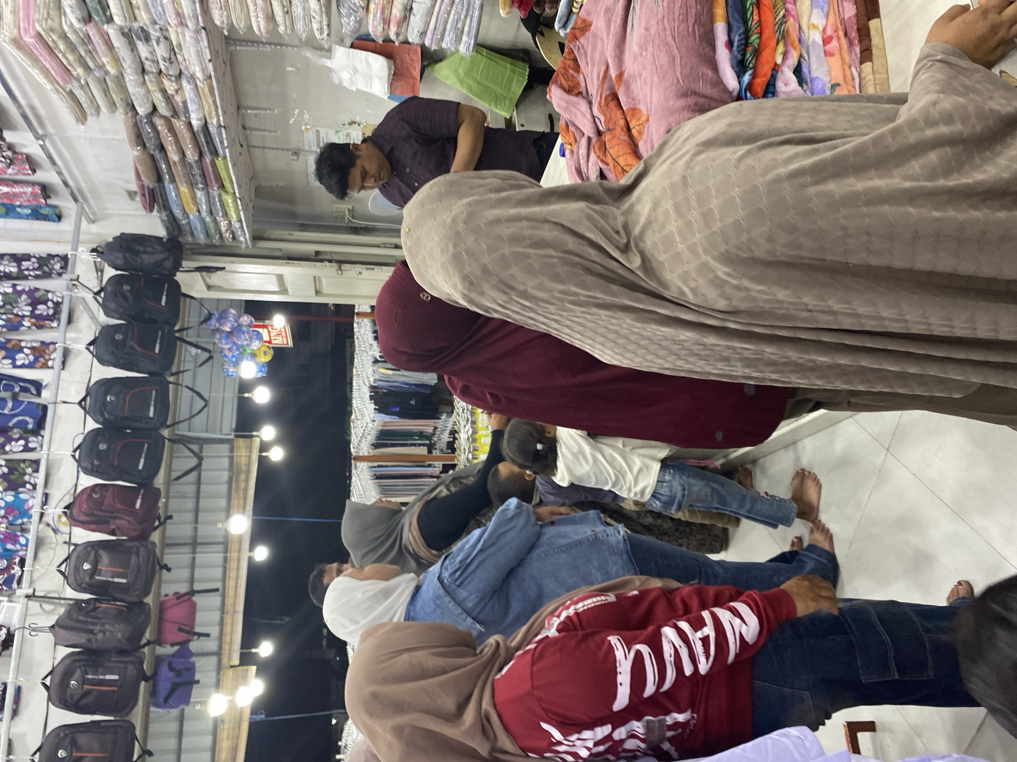 In Lamteumen Barat, Indonesia, a bustling marketplace scene unfolds within a store displaying various goods. The primary focus is on a group of individuals, predominantly women in hijabs, browsing through a selection of fabrics and bags. In the foreground, a person wearing a tan hijab stands facing away from the camera, with their arm resting on a pile of colorful blankets. To their left, another person in a maroon hijab is visible.
A child in a white top and blue jeans is seen standing near the center of the image, appearing to be engaged with the adults. Further back, a man in a dark shirt is attending to customers or organizing merchandise. The store's interior is densely packed with items. Rows of shelves are laden with folded fabrics, blankets, and other textiles. Hanging from above are numerous backpacks in various colors. The lighting suggests it may be evening or the store is dimly lit. The floor is tiled, and the overall impression is one of a busy retail environment.