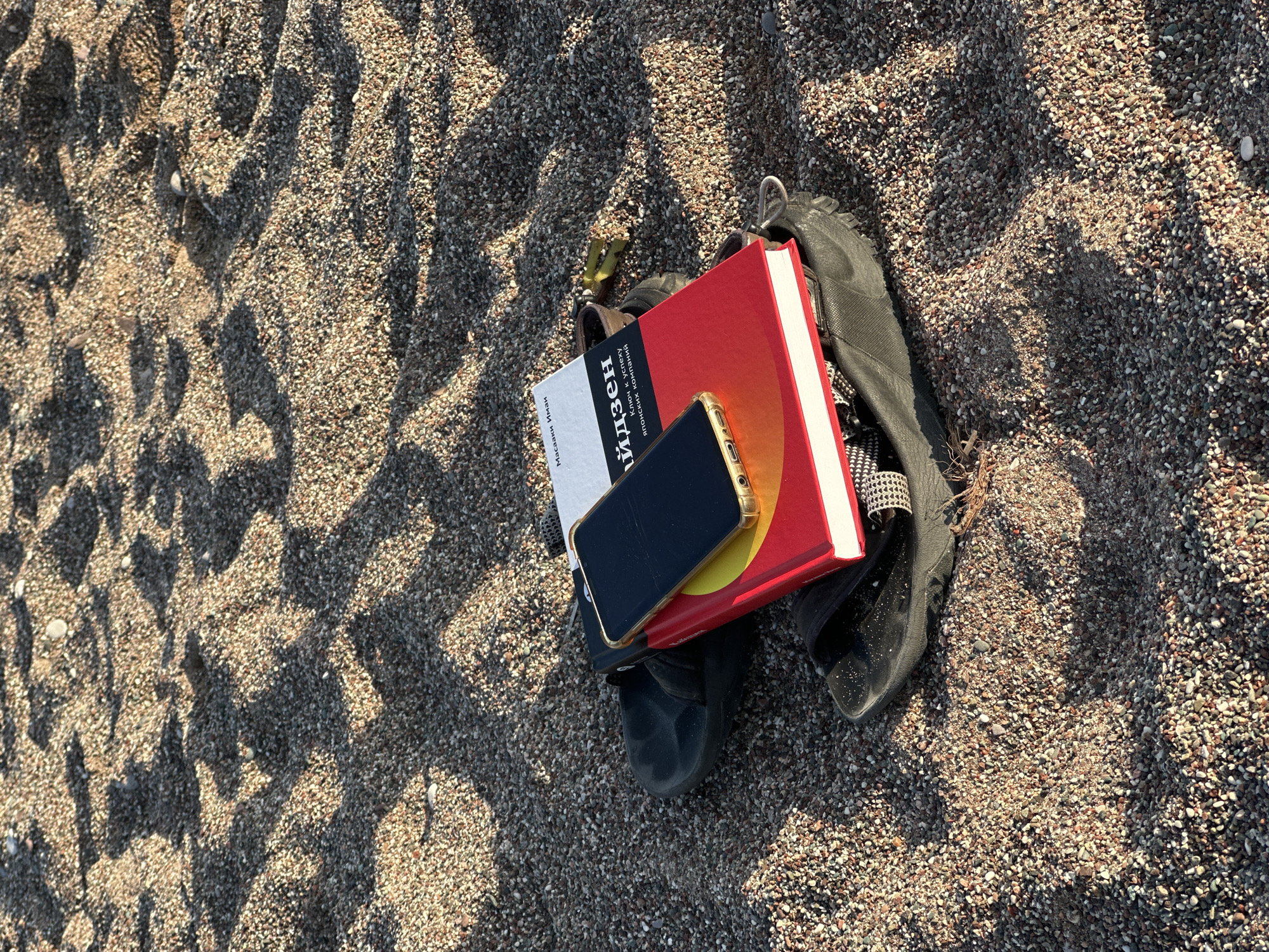 The image captures a close-up, high-angle view of personal items resting on a pebbly beach, likely in Bečići, Montenegro. The scene is illuminated by bright sunlight, casting long, distinct shadows across the textured surface, suggesting either early morning or late afternoon.
The primary subjects are a collection of objects arranged on the coarse, mixed-tone sand and pebbles. In the center, two books are stacked: a red-covered book underneath, and a smaller, white-covered book on top. A black smartphone with a prominent gold case is placed face-down on the white book. To the right of the books, a pair of black sandals or flip-flops is visible, and a small, metallic object (possibly a keychain or clip) is nestled among the items.
The white book's spine features visible Cyrillic text. The largest word is "ГЭЙДЗЕН", which is likely a transliteration of "Kaizen." Below it, smaller text reads "ЦИНЦОУ ХУНШИК" and "ИСКЭЙ ХОЙ" which appear to be names or publishing details. At the very bottom of the spine, "МАСАКИ ИМАИ" is visible, identifying the author Masaki Imai, known for books on Kaizen. No discernible text is visible on the red book.
The activity implied is one of leisure and relaxation, suggesting someone is enjoying a beach outing and has set aside their reading materials, phone, and footwear. The overall mood is calm and unhurried, characteristic of a vacation or day off by the sea.