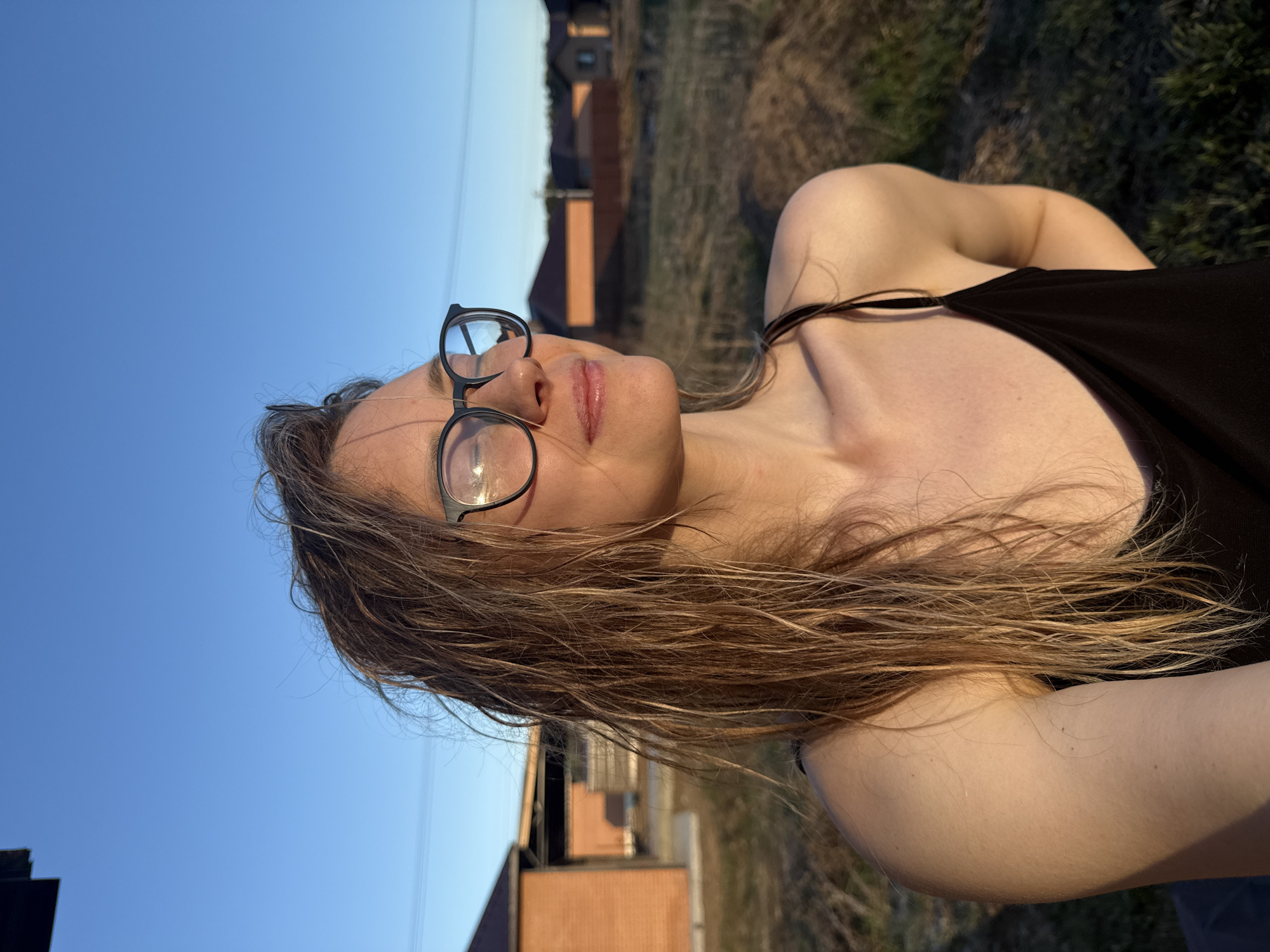 A young woman with long, light brown hair and wearing black-rimmed glasses poses outdoors. She wears a black tank top. The background features a grassy area, a blue sky, and buildings that appear to be residential. The location might be Chaikovsky, Russia, due to the building architecture. The image appears to be a selfie or photo taken at close range.