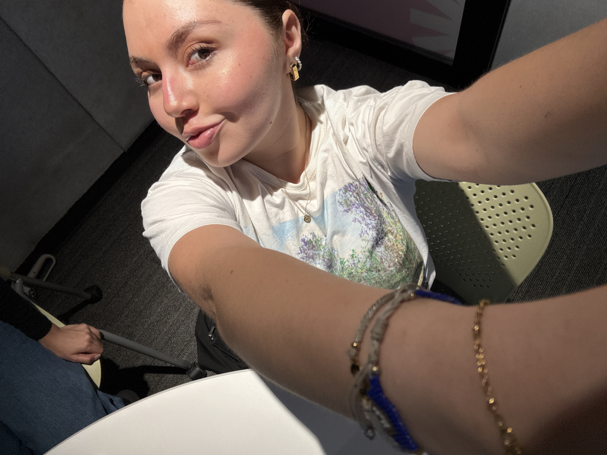 A fair-skinned young woman with dark hair, wearing a white t-shirt, gold earrings, and a gold necklace, is taking a selfie. She is indoors, seated at a white round table. A second person's legs are visible in the bottom left corner of the frame. The environment includes a chair, dark carpeting, and a partially visible window. The specific location in Mexico City, Mexico is not discernable from the image.