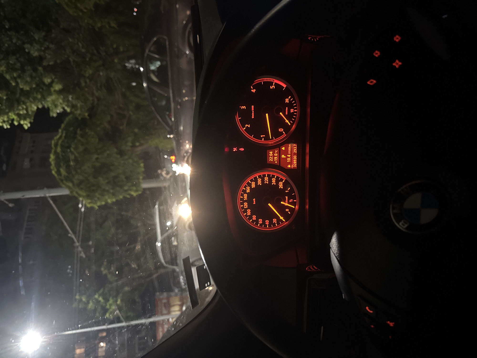The image depicts the dashboard of a BMW, presumably taken at night, while stationary. The instrument panel is illuminated in red, displaying the speedometer and tachometer. The speedometer reads approximately 20 km/h, and the tachometer is slightly above 0. The odometer shows a reading of 328930 km, and a consumption rate of 10.4 l/100km. The time is 22:04, and the temperature is +19.0°C. Through the windshield, a street scene with trees and buildings is visible, indicating an urban environment. The BMW logo on the steering wheel is partially visible. The image is slightly tilted. The location is Stara Zagora, Bulgaria, is speculative and not directly verifiable from the image alone.