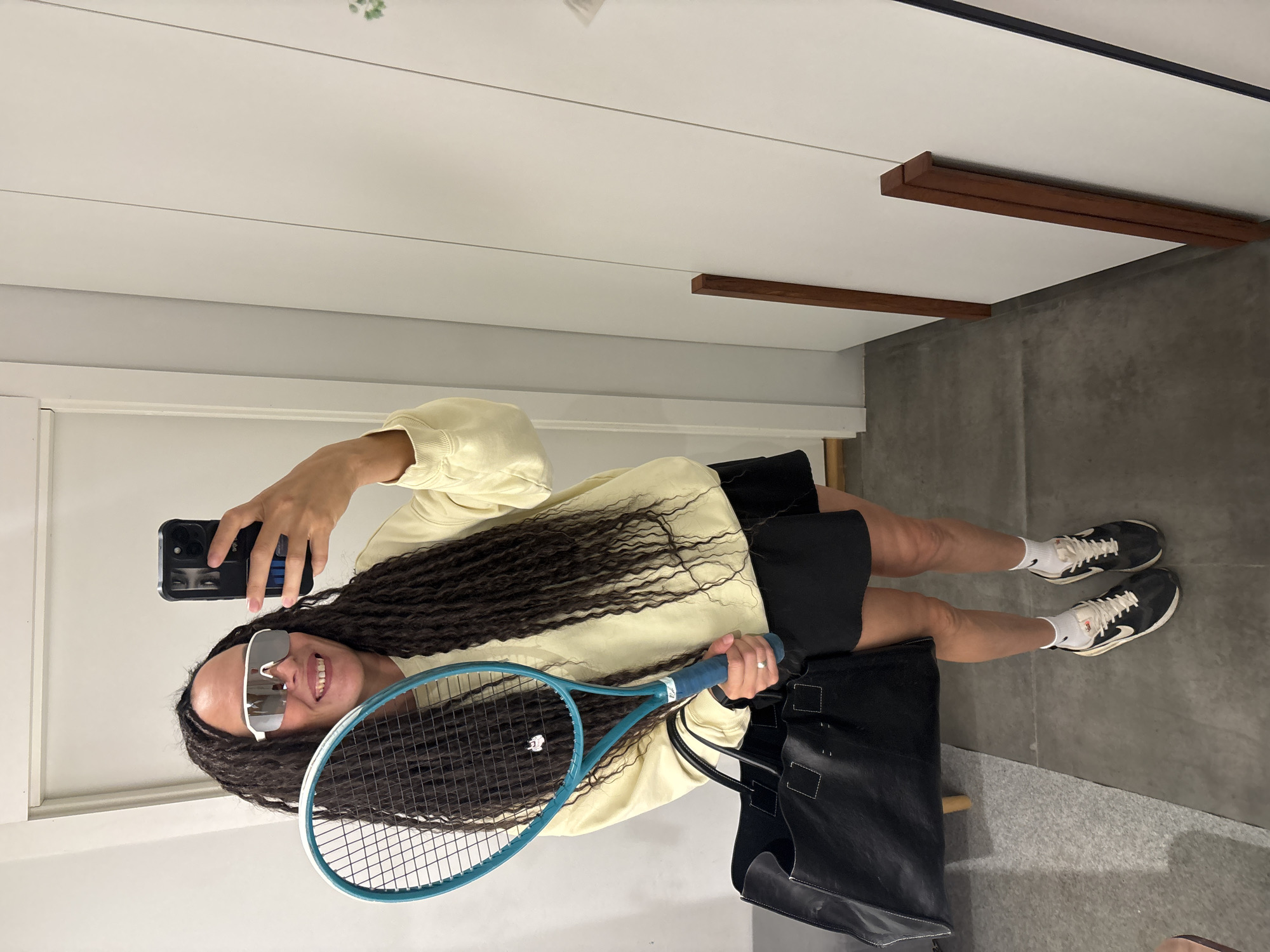 A full-body, vertically oriented (rotated 90 degrees clockwise from original capture) image shows a person taking a mirror selfie indoors. The individual, appearing female, has long, dark, curly hair and is smiling broadly, revealing teeth. She wears large, reflective, white-framed sunglasses.
She is dressed in a light yellow long-sleeved sweatshirt and black shorts. On her feet are black sneakers with white soles and laces, paired with white athletic socks that display the "Nike" swoosh logo.
In her right hand, she holds a black smartphone with a textured black case, angled towards her for the selfie. The reflection in the phone screen shows a partial view of her eyes. In her left hand, she holds a blue or turquoise tennis racket. A large black tote bag with white stitching details rests on the floor beside her.
The background suggests a modern indoor setting. The ceiling is white with thin horizontal lines and features decorative wooden beam accents. The walls are predominantly white, with a section of dark gray, possibly concrete, visible on the right. The floor is a light gray. The overall ambiance appears bright and well-lit, indicative of daytime. The location context provided is Chaikovsky, Russia.