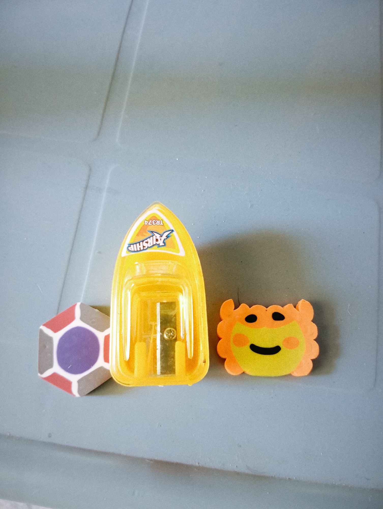 Three small stationery items are arranged on a grey surface. On the left is a hexagonal eraser with a geometric pattern. The pattern consists of a purple circle in the center, surrounded by alternating red and grey hexagonal shapes. To the right of the eraser is a bright yellow pencil sharpener. The sharpener is shaped like a small boat or airship, with a blue and yellow logo on its upper surface. Visible text on the logo reads "AIRSHIP" and "TR374". The pencil sharpening mechanism is visible at the front of the sharpener. To the right of the pencil sharpener is a small, orange and yellow eraser shaped like a cartoon lion's face. The lion has black dots for eyes, pink circles for cheeks, and a black curved line for a mouth. The background surface appears to be a plastic or laminate material with some subtle linear indentations, suggesting it might be a desk or tray. The lighting is even and appears to be from an overhead source, casting soft shadows. There are no people or animals visible, and the scene is static, with no activity taking place.