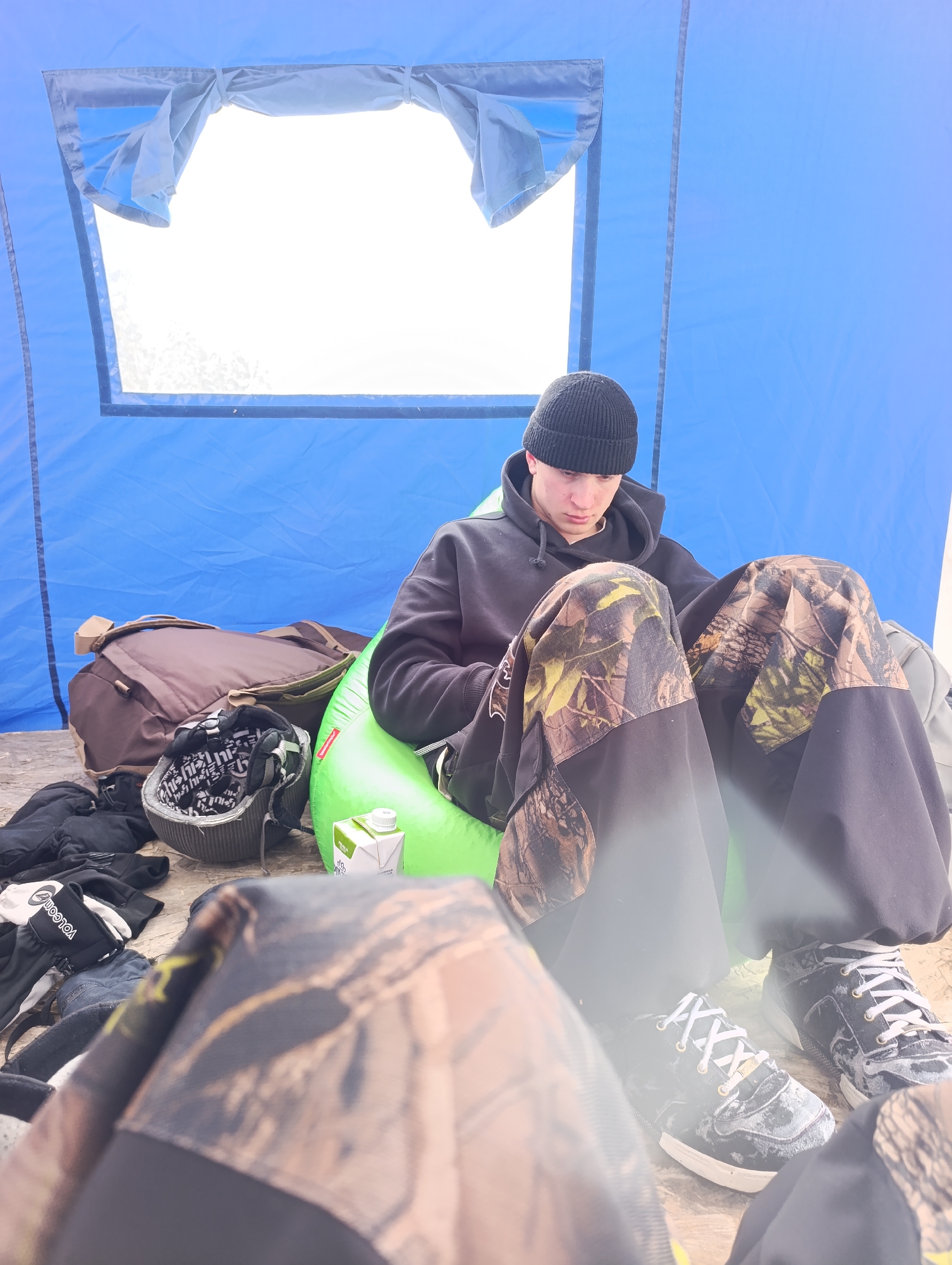 A young man sits inside a blue tent in what appears to be an indoor or semi-indoor setting. He is wearing a black beanie, a black hoodie, and camouflage-patterned pants with black portions. He is sitting on a green inflatable seat. Beside him, there is a brown backpack, a helmet, gloves, and a small carton of juice. A window in the tent shows a bright, overexposed outdoor scene. The scene is cluttered with clothing and gear. There is an additional set of camouflage pants in the foreground obstructing part of the view. The photograph does not contain enough information to determine if it was taken in the city of Krasnoyarsk, Russia.