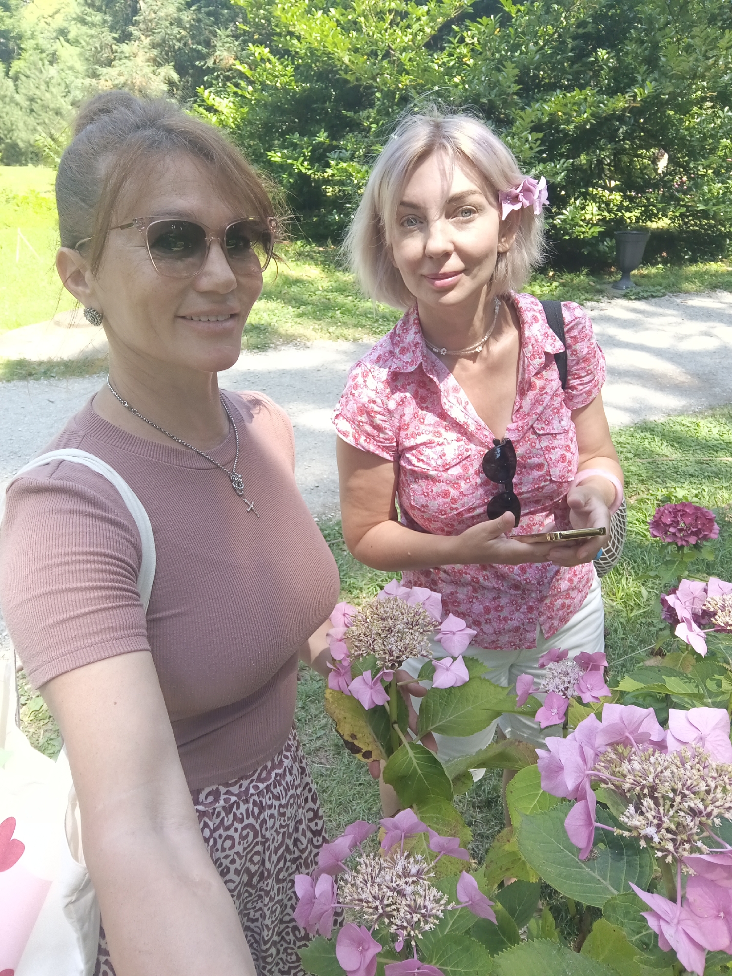 Two women are posing for a selfie outdoors, surrounded by greenery and pink hydrangeas. The woman on the left has her hair pulled up in a bun and is wearing sunglasses and a mauve ribbed t-shirt. She is also wearing a leopard print skirt and a silver necklace with a cross pendant. The woman on the right has short blonde hair with a pink flower tucked behind her ear. She is wearing a pink floral print shirt and white shorts, with a silver necklace. She is holding a smartphone in her right hand. The background consists of trees and a gravel path. The location is identified as Bestuzhevskoye, Russia.
