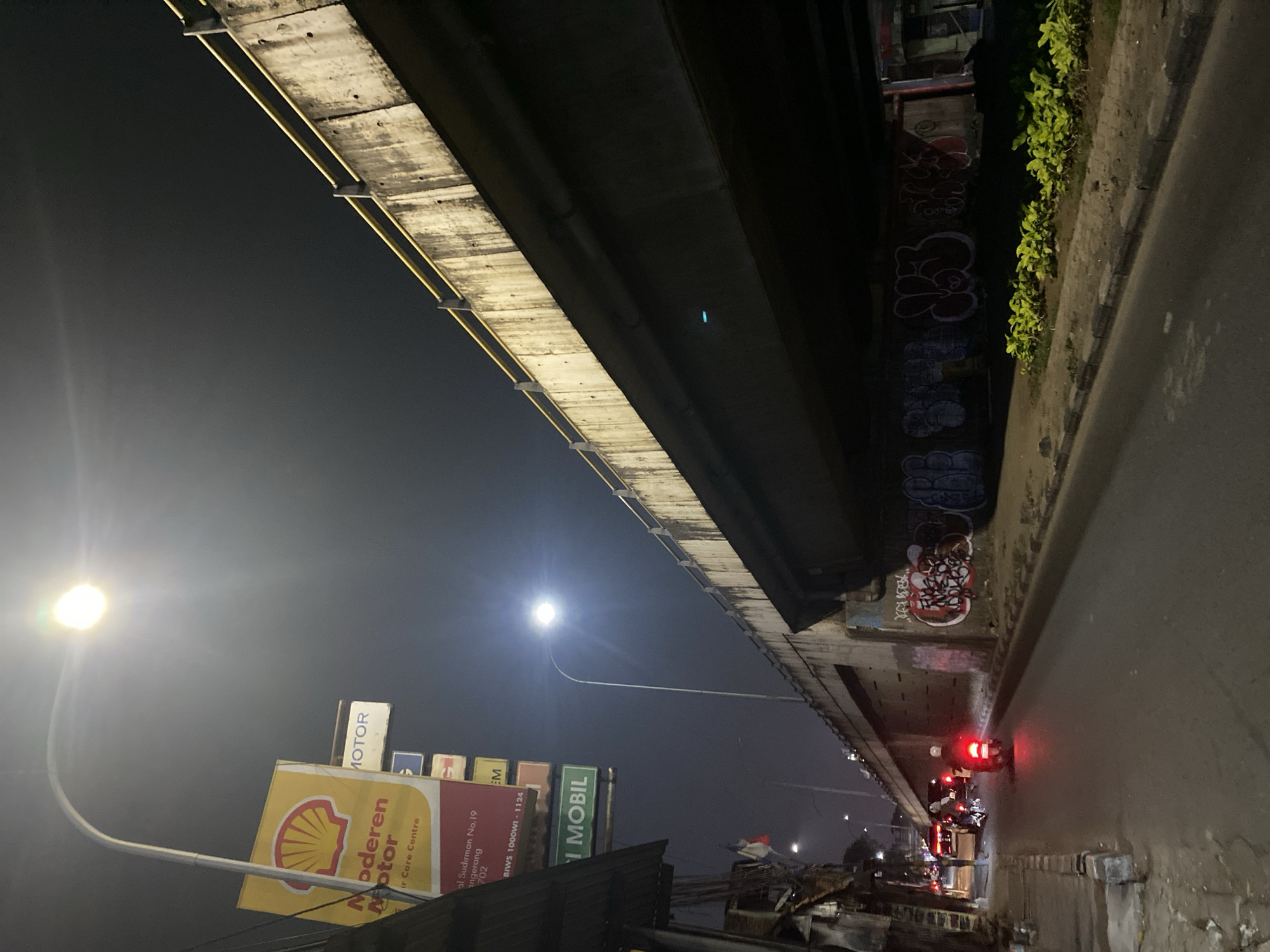 An outdoor, low-angle, wide shot captures a nighttime scene in Tangerang, Indonesia, beneath an overpass. The sky is dark and clear. A bright street lamp illuminates the left side of the frame, casting a strong glow. To the left, a yellow Shell sign with a red scallop logo is visible, reading "Moderen Motor" and "Car Care Centre." Below it, a red sign displays "Jl. Sudirman No. 19 Tangerang 15112." Further down, a green sign reads "MOBIL." A few buildings with metal roofs and some wires are present on the far left.
The concrete overpass dominates the upper right portion of the image, its textured surface appearing aged and streaked. A yellow railing runs along its edge. The underside of the overpass is dark, with graffiti visible on the concrete pillars and walls. The graffiti consists of various stylized lettering and designs in red, white, and blue.
A paved road runs from the bottom center towards the background. Several people on motorcycles are visible on the road. One motorcycle in the foreground has its red taillight illuminated, casting a red glow on the road. Other motorcycles and a car are further back, their headlights and taillights visible. A strip of small, bright green plants lines the edge of the road beneath the overpass. The scene suggests a busy urban environment at night.