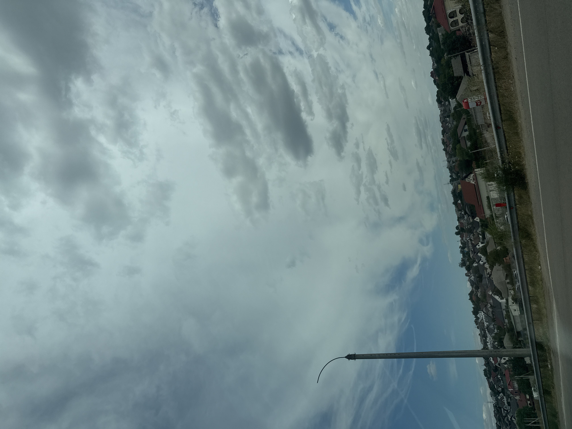 The image shows a view of a residential area on a hillside, likely in Băcioi, Moldova, under a cloudy sky. In the foreground, there is a paved road. A metal barrier runs along the edge of the road, separating it from a grassy area with dry vegetation. A tall, gray utility pole with a curved arm is prominently positioned on the left side of the frame. The hillside is densely populated with houses of various sizes and architectural styles, many with red or dark-colored roofs. Trees and vegetation are scattered throughout the residential area. The sky is filled with a mix of gray and white clouds, with some patches of blue sky visible, suggesting variable weather conditions. There are no people or vehicles clearly visible in the image. The overall impression is that of a suburban or semi-rural landscape under an overcast sky.