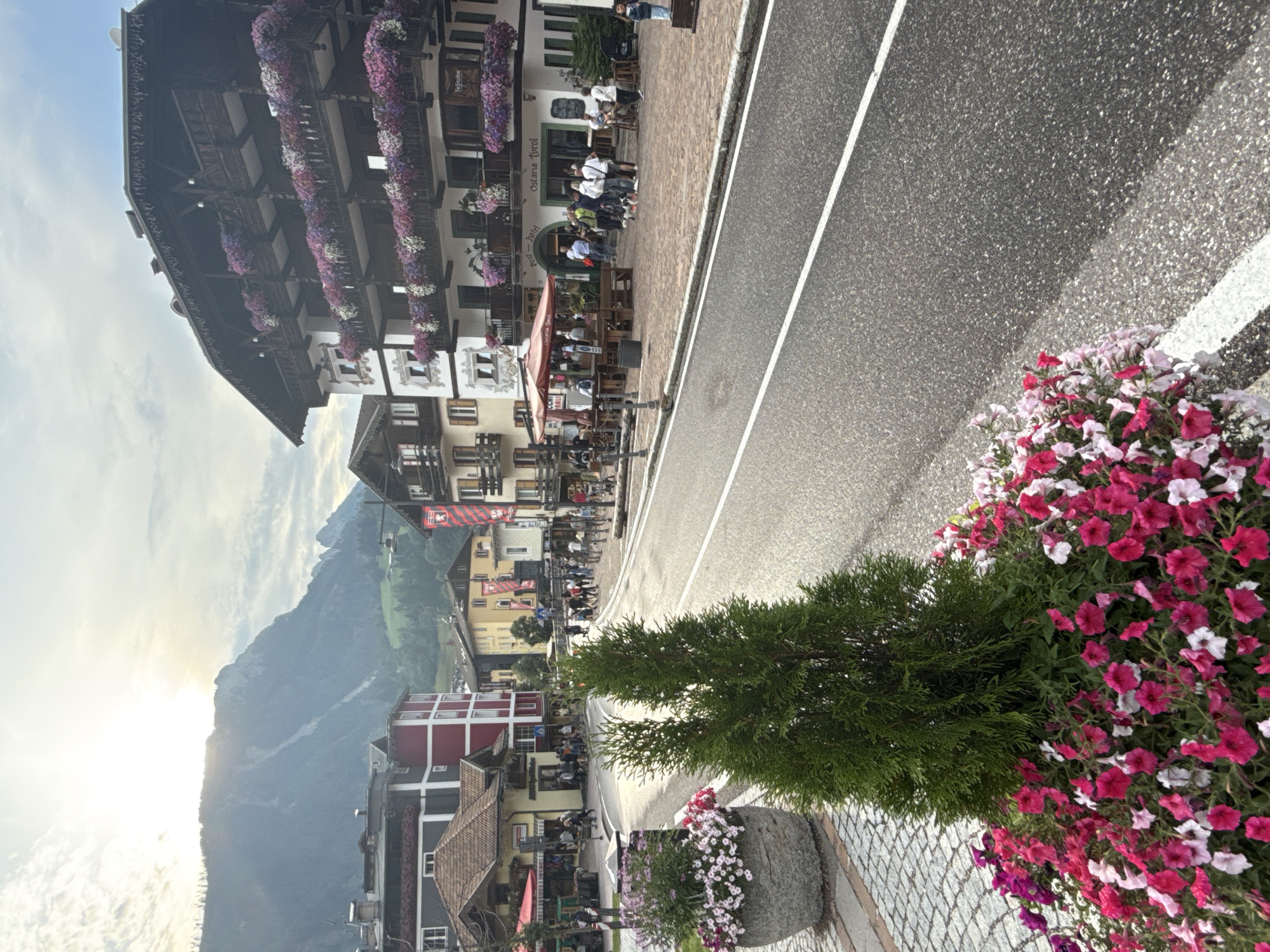 The image depicts a street scene in Moena, Italy, a town in the Dolomites region, characterized by its alpine architecture and mountainous backdrop. In the foreground, two large stone planters overflow with vibrant pink and white petunias and a small evergreen tree. A cobblestone sidewalk runs along the left side of the street, leading to a paved road with clear white lane markings.

On the right side of the street, several buildings with traditional Tyrolean architectural styles are visible. One prominent building, a hotel with balconies adorned with colorful flowers, features signs indicating "Post Hotel" and "Osteria Tyrol." People are gathered on the sidewalk outside this establishment, some standing in groups, others walking, and a few appear to be seated at outdoor tables under a red umbrella. Several dogs are also present, some on leashes and one seemingly interacting with a person.

Further down the street, more buildings line the road, some painted in muted yellow and red tones, interspersed with shops and cafes. Banners with red and white motifs are displayed on poles. In the background, a majestic mountain range is partially obscured by the sun, which shines brightly through a cloudy sky, casting a soft light on the scene. The overall impression is of a lively, picturesque town square on a pleasant day.