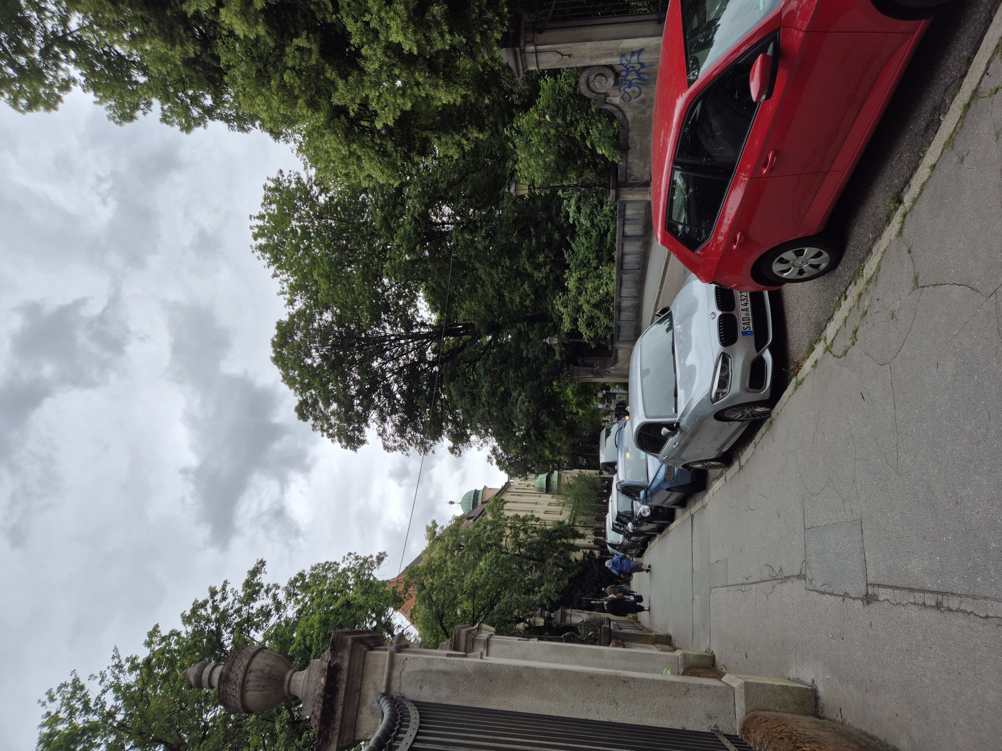 The image depicts a street scene in Regensburg, Germany, on a cloudy day. A sidewalk runs along the left side of the frame, bordered by a stone wall and a black metal fence. Several people are walking on the sidewalk, heading away from the camera. A row of parked cars lines the right side of the street, including a red hatchback in the foreground, a silver sedan, and several other vehicles further down. Behind the cars, lush green trees form a dense canopy, and a building with an orange tiled roof and green domes is visible in the background. The atmosphere is overcast, with dramatic clouds filling the sky.