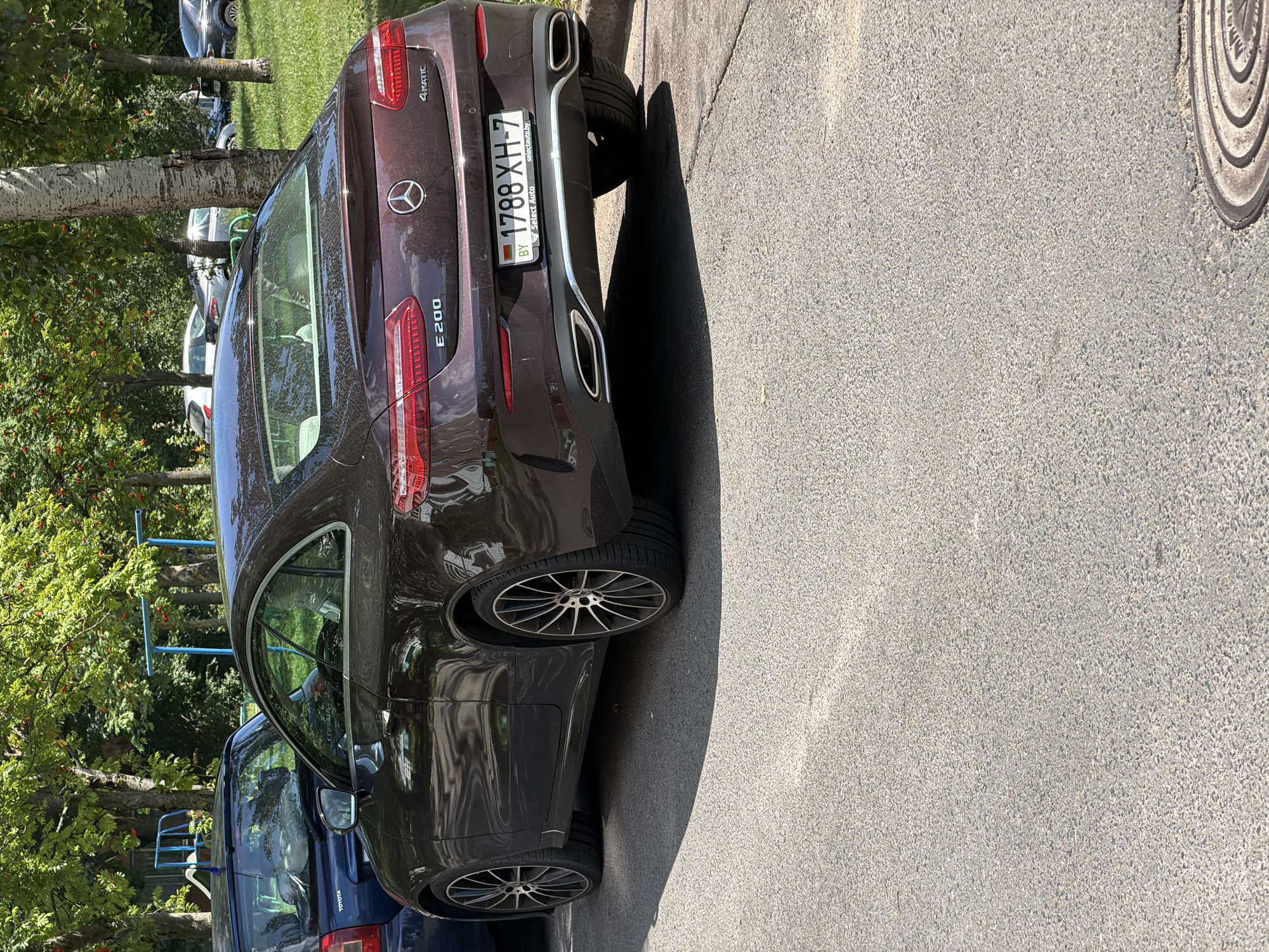 A dark red Mercedes-Benz E 200 4Matic coupe is parked on a paved road in Minsk, Belarus. The car is facing left, and its rear is visible. The license plate reads "1788 XH-7". The car is parked next to a blue Skoda Octavia. In the background, trees with green foliage and some red berries are visible, along with a children's playground with blue bars and a slide. Other cars are parked in the distance. The scene appears to be a residential area on a sunny day.